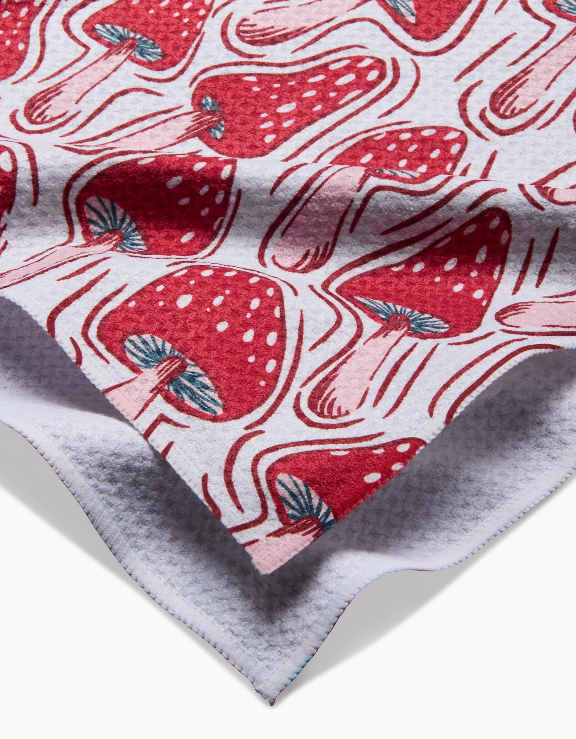 A close-up of Hufton Studios Christmas Mushrooms tea towel shows its soft Geoweave fabric, adorned with red mushrooms featuring white spots and teal stems on a light background. The slightly folded edge reveals the textured underside.