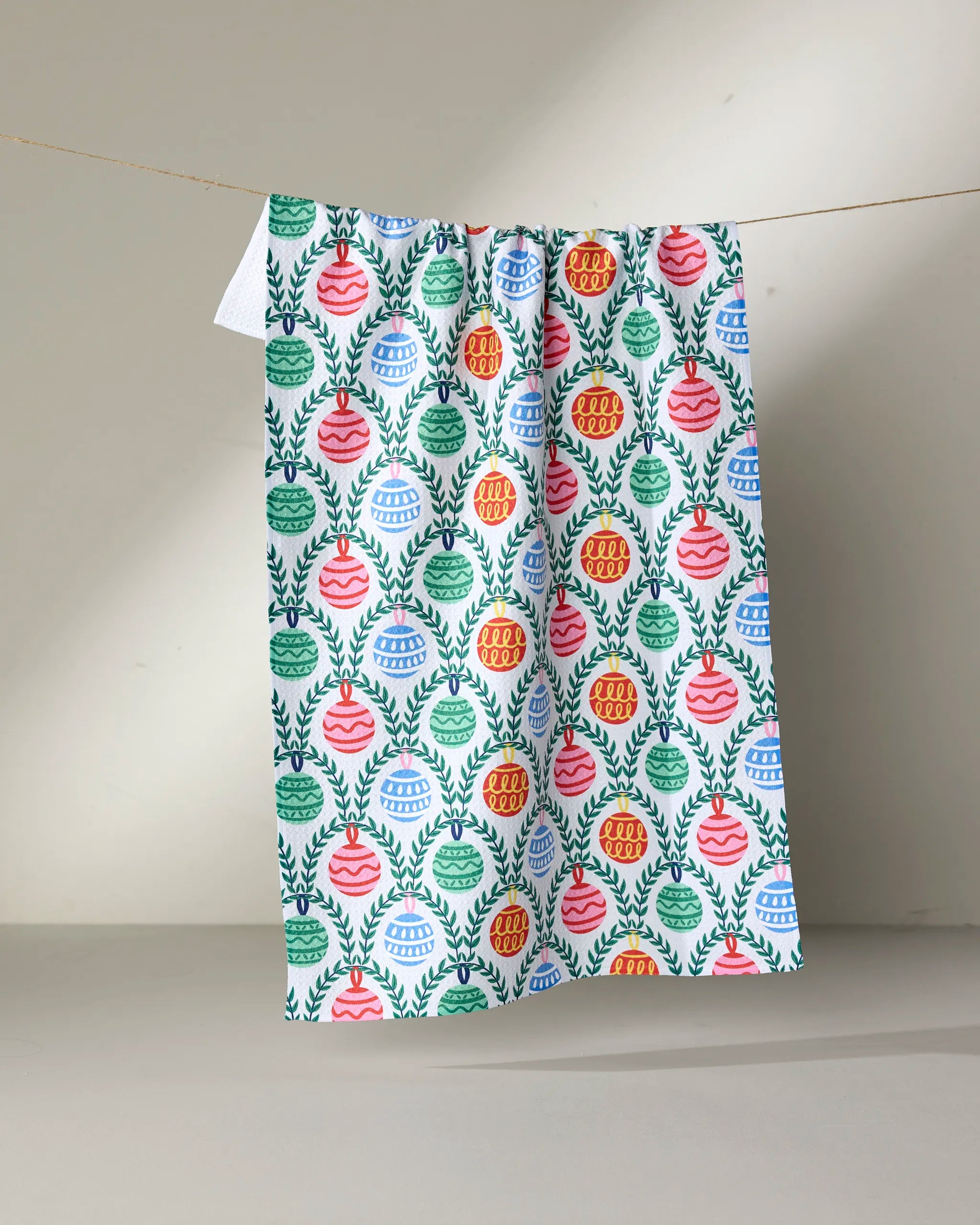 The Christmas Ornaments tea towel by Ceyda Alasar features rows of red, green, blue, and orange ornaments with leafy branches, displayed against a neutral background.