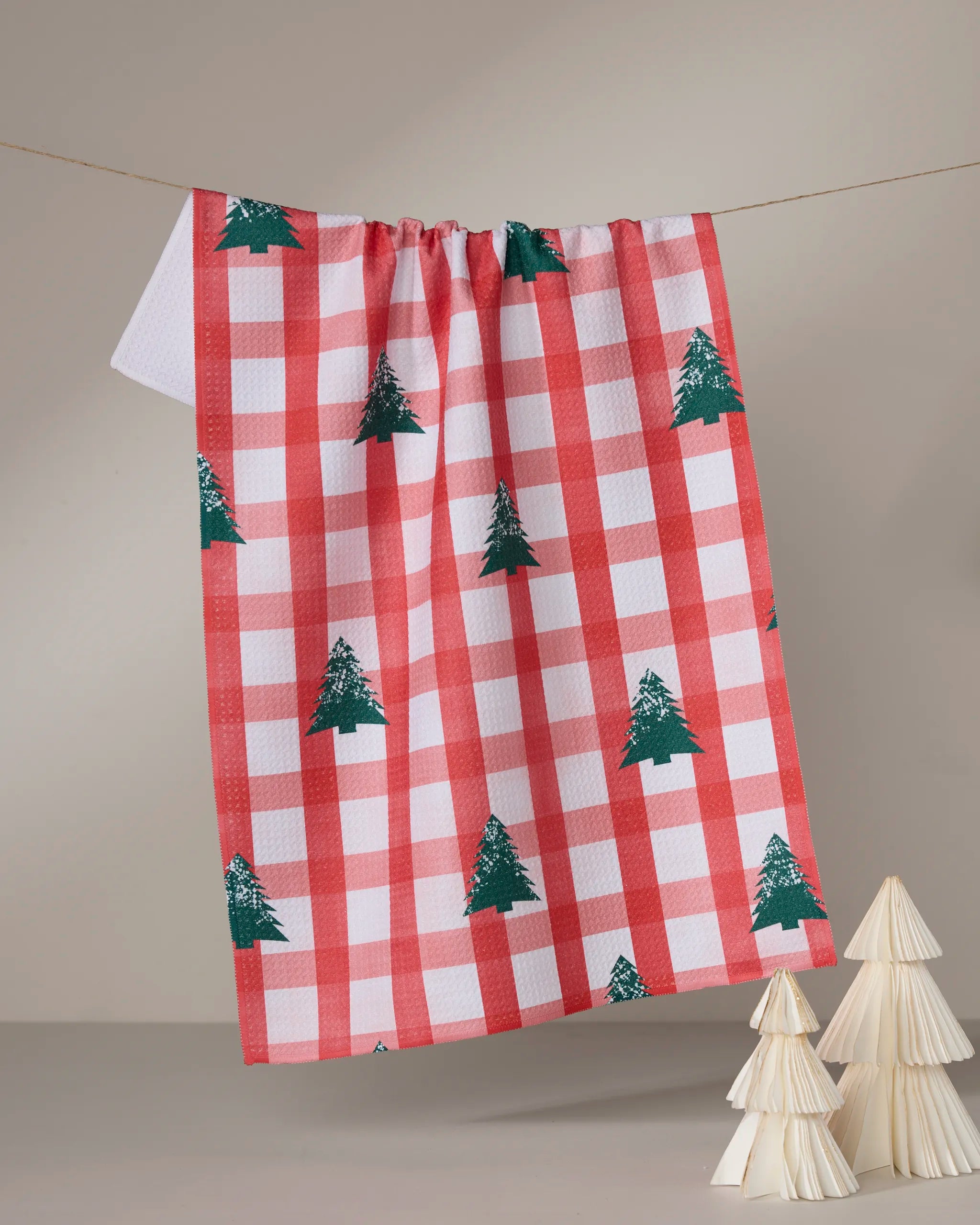 The Geometry Christmas Pines tea towel, featuring red and white checks with green Christmas trees, hangs on a string. Below, two small decorative paper trees rest against a neutral background.