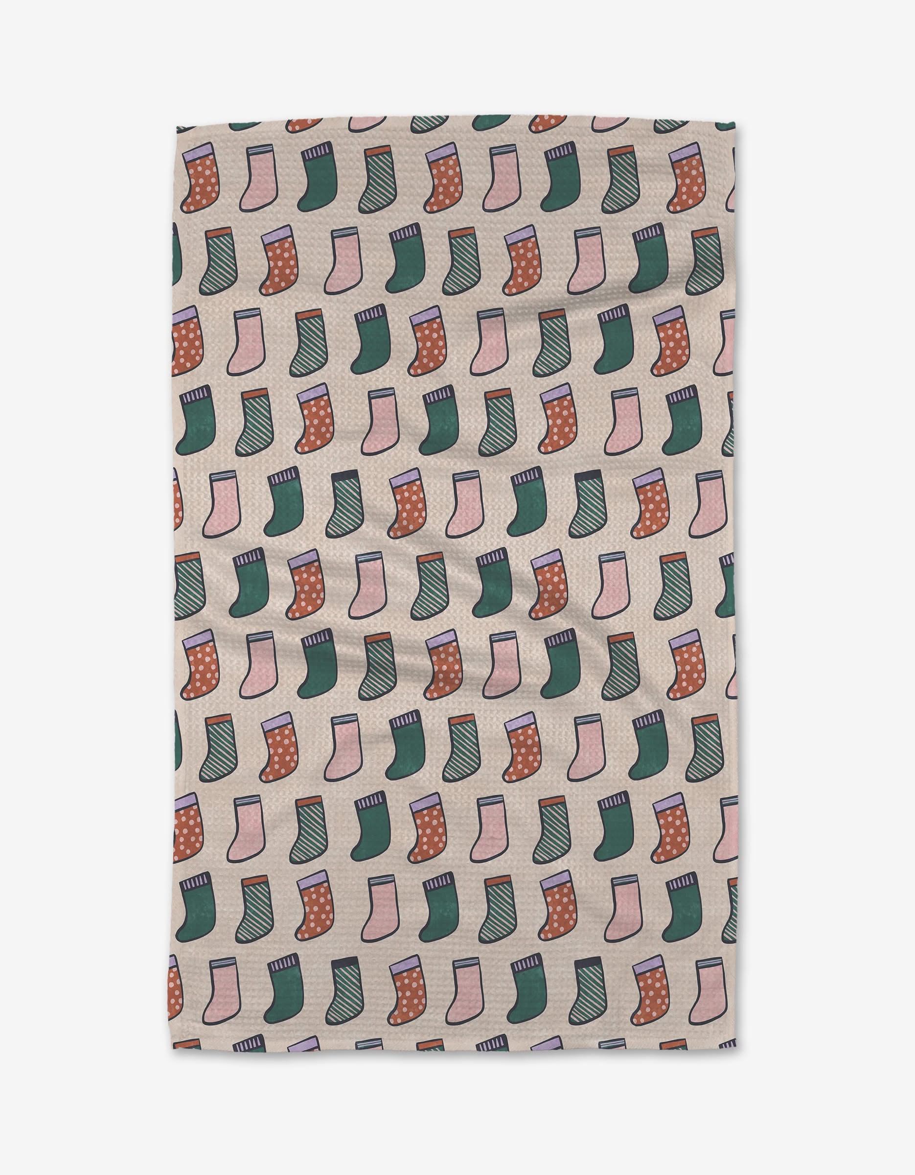 The Christmas Stockings kitchen towel by Claire Iglesias features rows of illustrated green, red, and pink stockings with festive patterns on a light beige background.