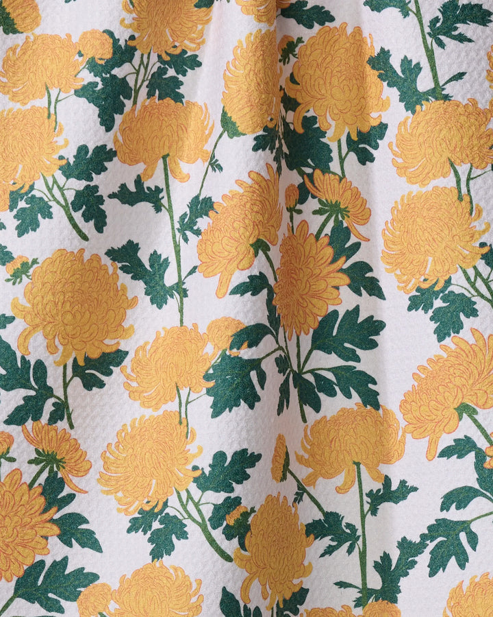 Yellow chrysanthemums with green leaves adorn textured white fabric, accented by a central crease that creates soft folds and shadows. The Vaishali Design Studio "Chrysanthemum" tea towel is crafted from recycled materials.