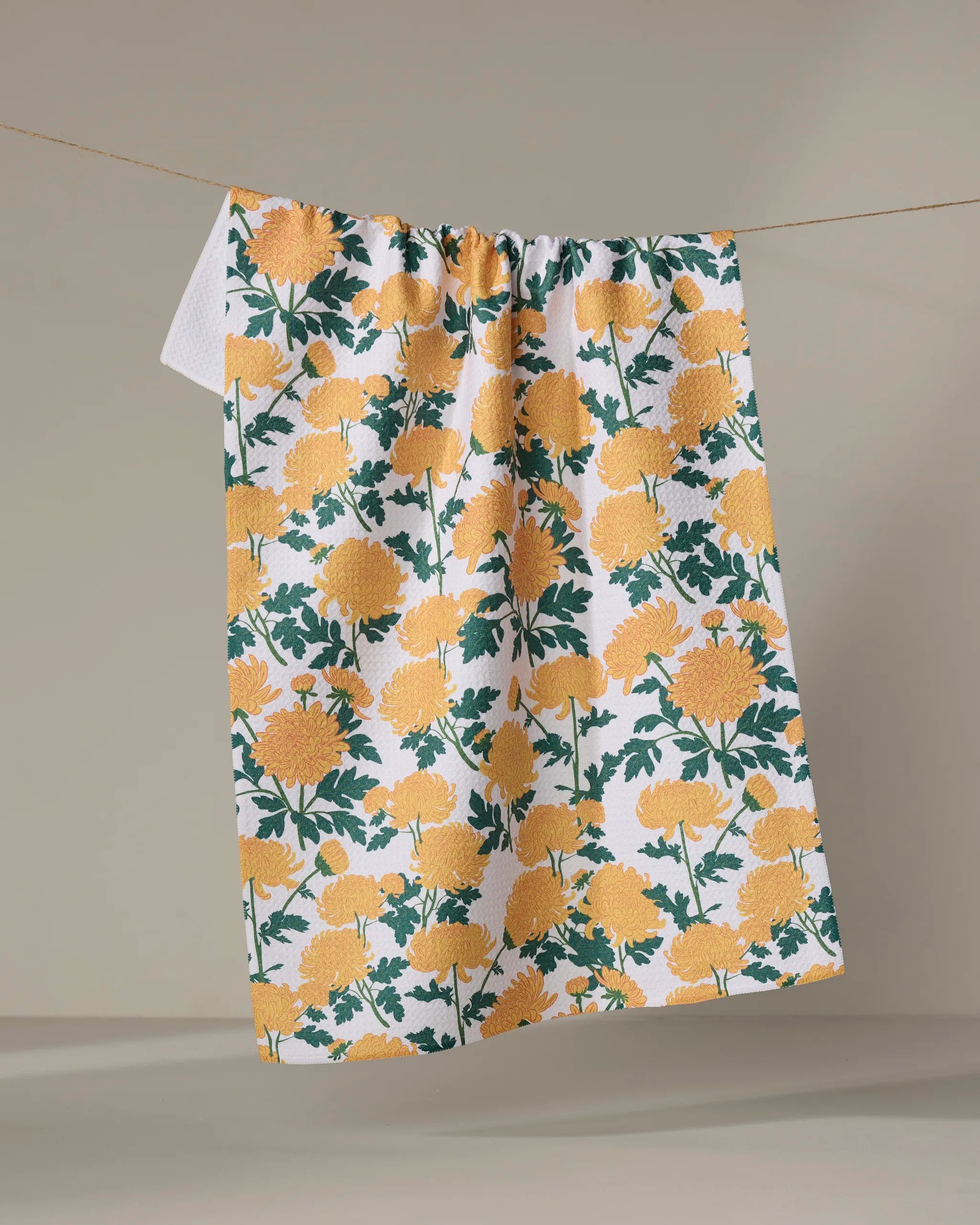 The "Chrysanthemum" tea towel by Vaishali Design Studio features a yellow floral pattern and green leaves on white, crafted from recycled materials for an eco-friendly touch, shown hanging on a clothesline against a neutral background.