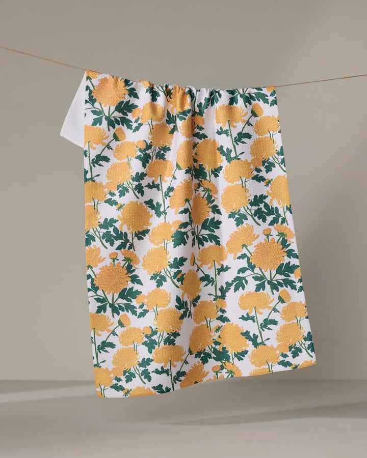 The "Chrysanthemum" tea towel by Vaishali Design Studio features a yellow floral pattern and green leaves on white, crafted from recycled materials for an eco-friendly touch, shown hanging on a clothesline against a neutral background.