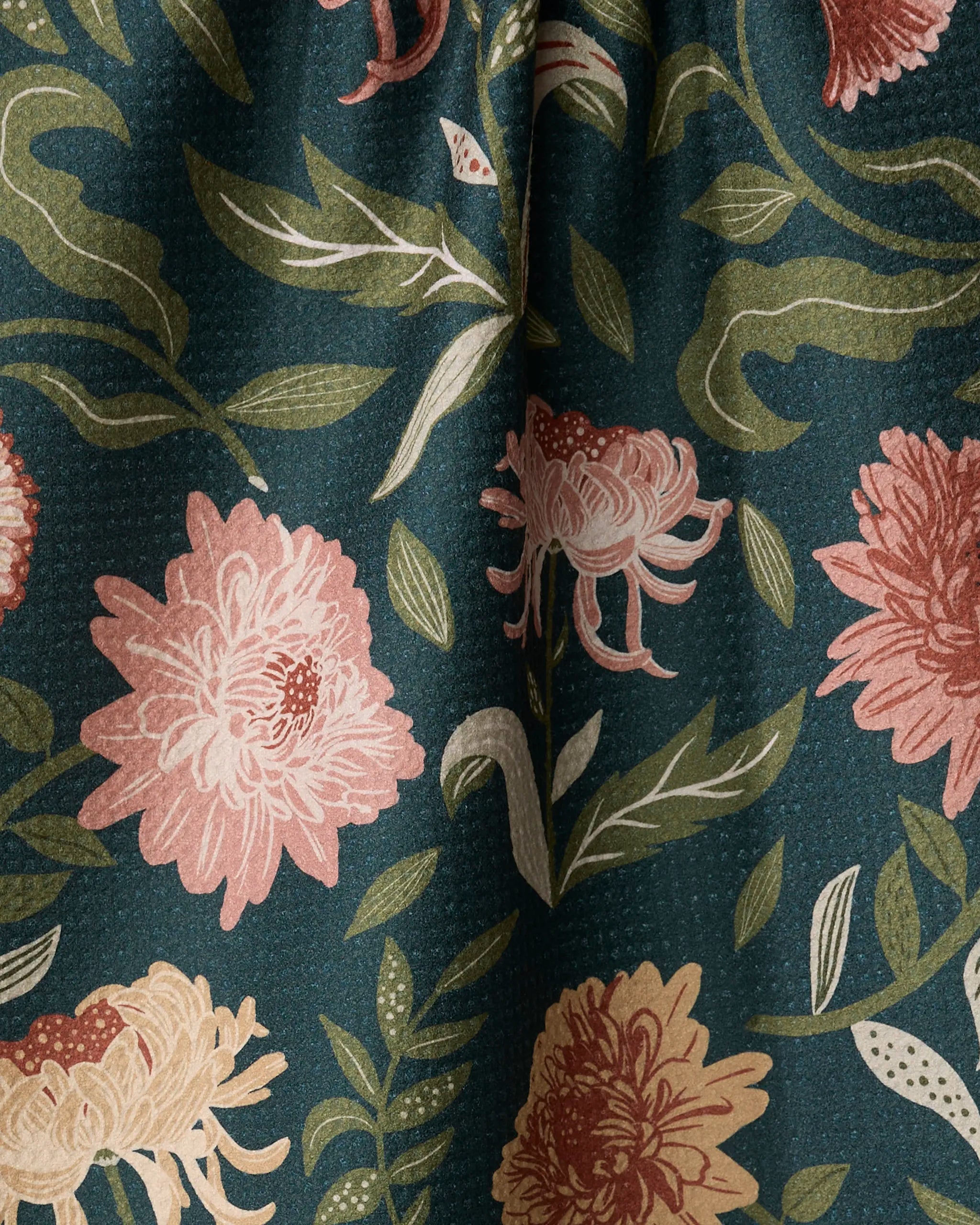 A close-up of the Chrysanthemums In November tea towel by Danelys Sidron, showing ultra-absorbent, textured fabric with large pink and beige chrysanthemums, green leaves, and stems on a dark green background.