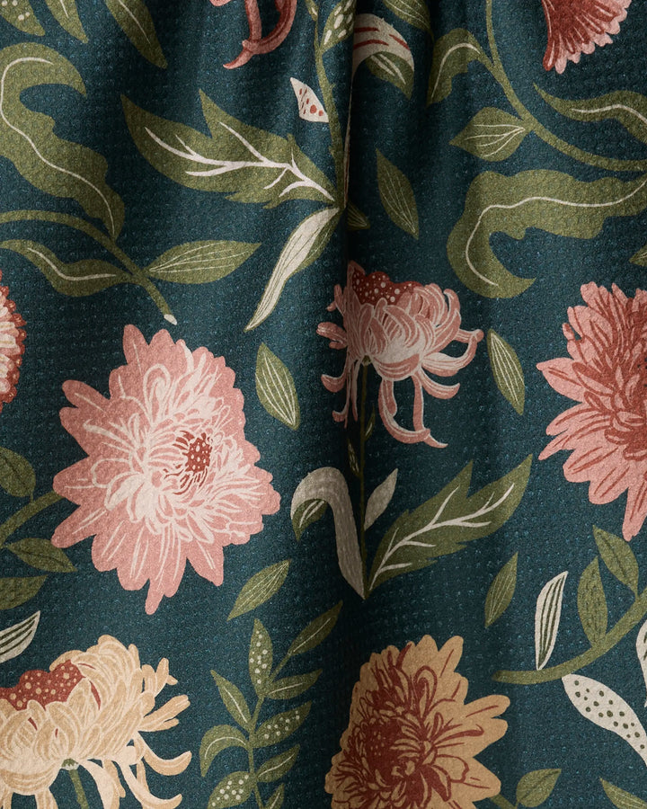 A close-up of the Chrysanthemums In November tea towel by Danelys Sidron, showing ultra-absorbent, textured fabric with large pink and beige chrysanthemums, green leaves, and stems on a dark green background.