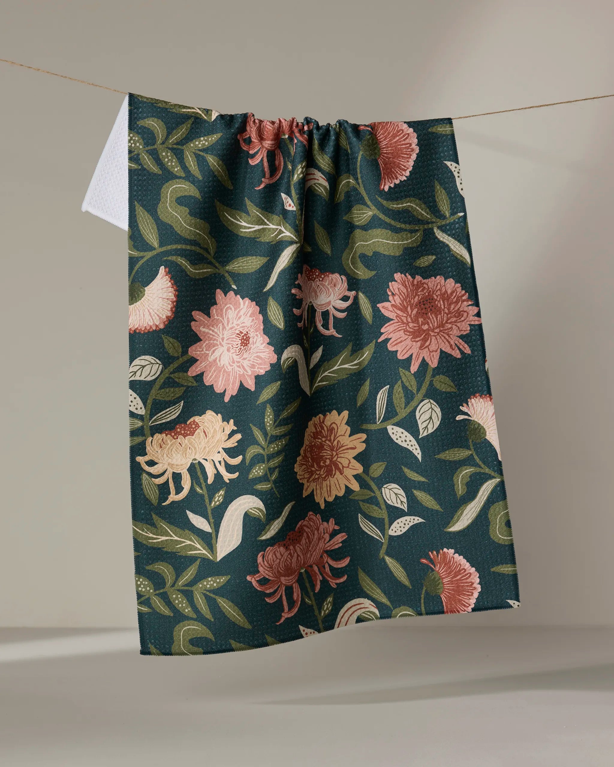The "Chrysanthemums In November" tea towel by Danelys Sidron, ultra-absorbent and featuring a dark green base with pink and cream floral patterns, hangs on a clothesline against a light background.