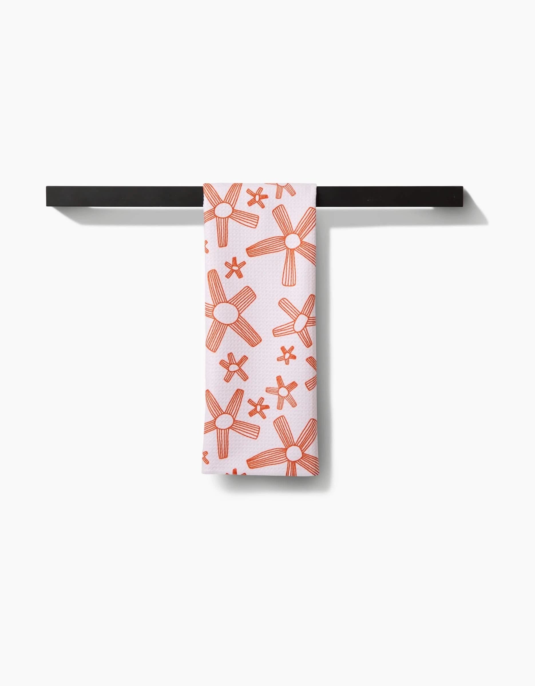 The Chunky Striped Flower tea towel by Coit Creative, crafted from recycled materials, features an orange floral pattern on white and is draped over a black towel bar against a white background.