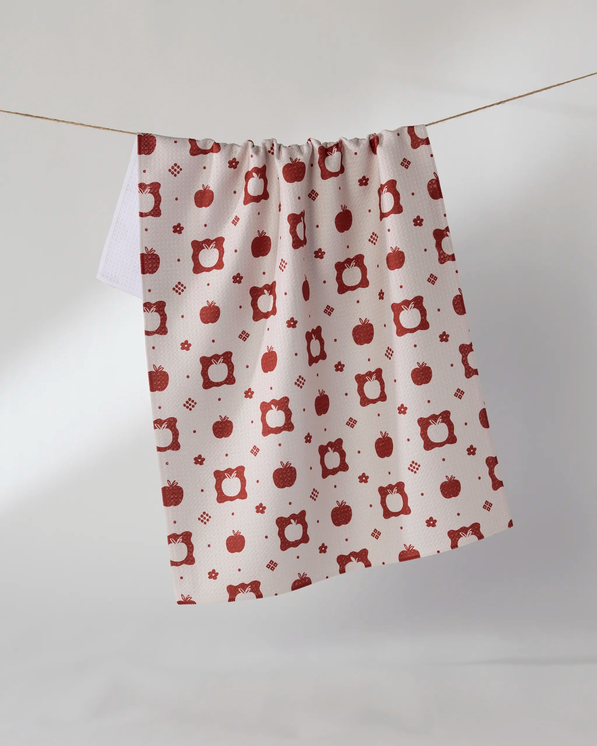 The Geometry Cider Mill tea towel, crafted from recycled materials, features a red apple and geometric design and hangs on a string against a plain light background.