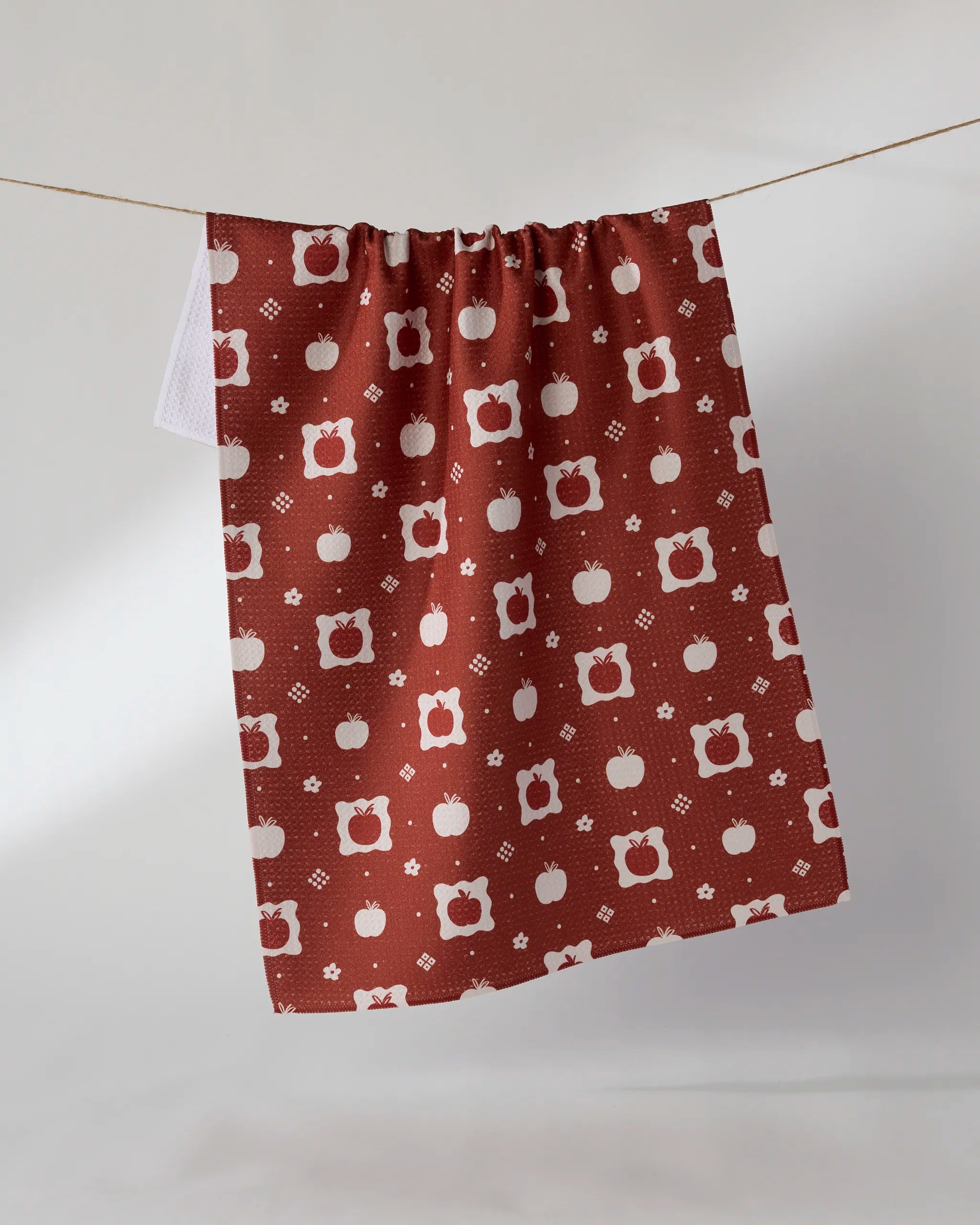 The Geometry "Cider Mill" tea towel, made from rust-colored Geoweave fabric and featuring a white apple and floral pattern, hangs on a string against a light background.