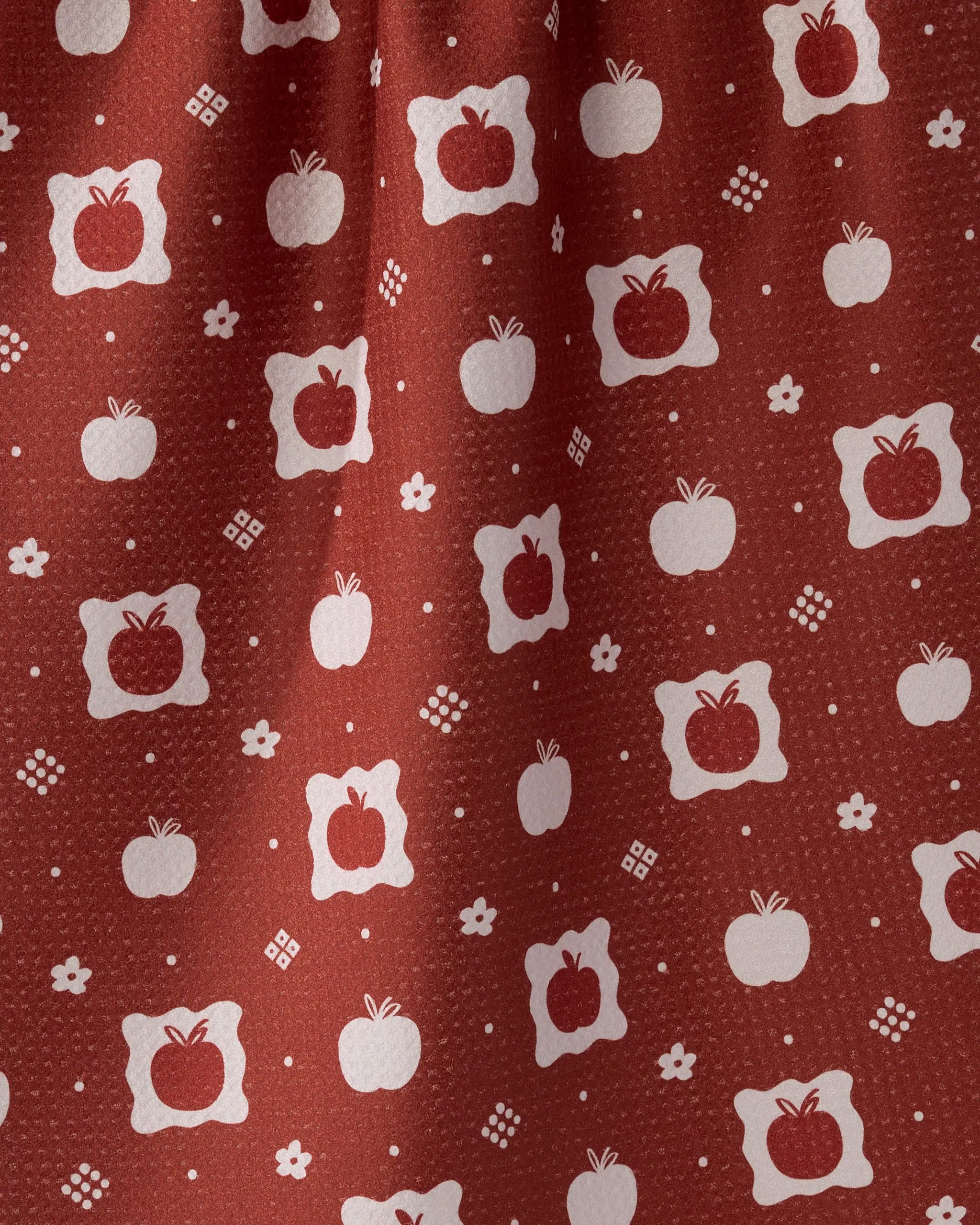 A close-up of Geometry's Cider Mill Geoweave tea towel, featuring white apples inside scalloped squares and dotted geometric accents on red, with a subtly textured surface and soft folds.