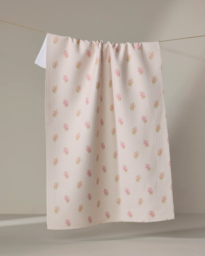 The Cindy tea towel by Kelsi Fullmer features a cream color, subtle textured pattern, and small pink flower prints. Made from recycled materials, it hangs neatly over a clothesline against a neutral background.