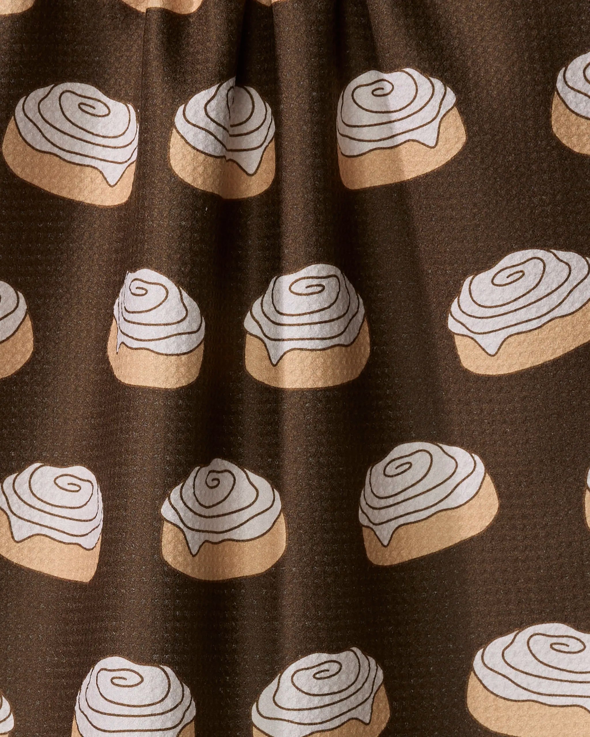 The "Cinnamon Girl" tea towel by Purely Kaylie features a brown hue with illustrated cinnamon rolls topped with white icing spirals. Made from recycled materials, it’s ultra-absorbent with a textured surface and gentle folds.
