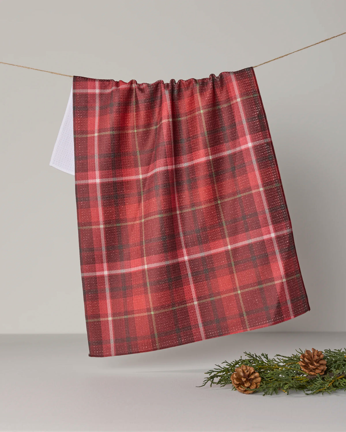 A Geometry Cinnamon Spice dish towel, featuring a red and green plaid design and highly absorbent fabric, hangs on a line against a plain background. Pinecones and evergreen sprigs are arranged on the surface below.