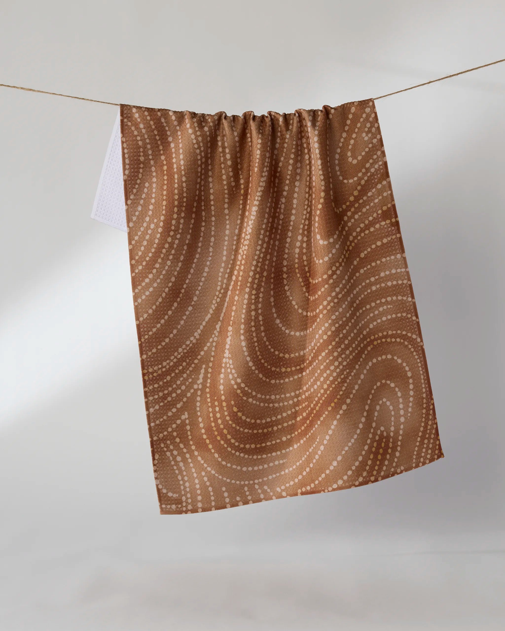The Geometry "Cinnamon Swirl" tea towel, crafted from recycled materials, displays wavy white dotted lines on brown and hangs on a string against a plain light background.