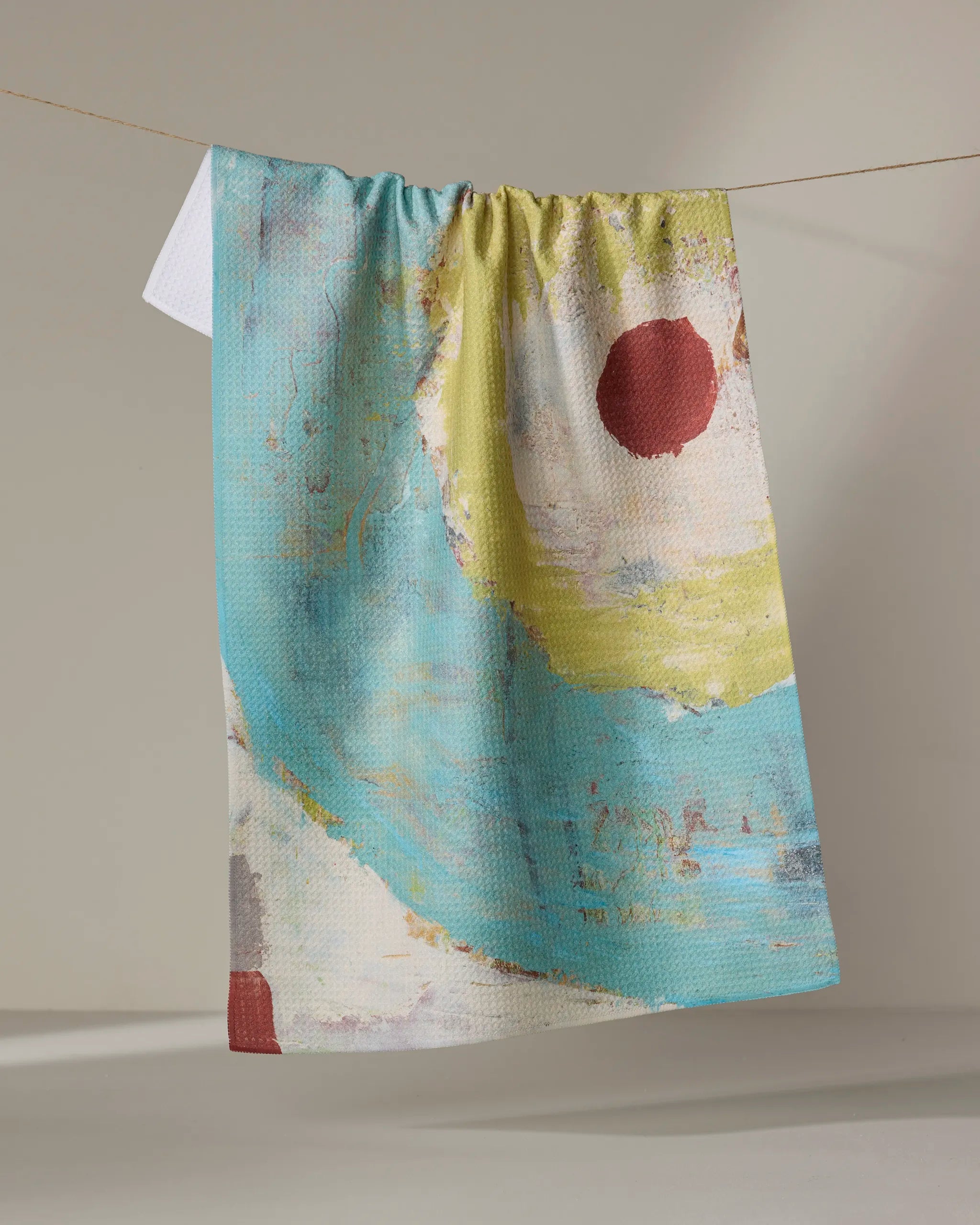 The "Circle Squared" kitchen towel by Katherine Berkeley features abstract blue, yellow, and white art with a bold red circle. Made from recycled materials, it hangs on a string against a neutral background.