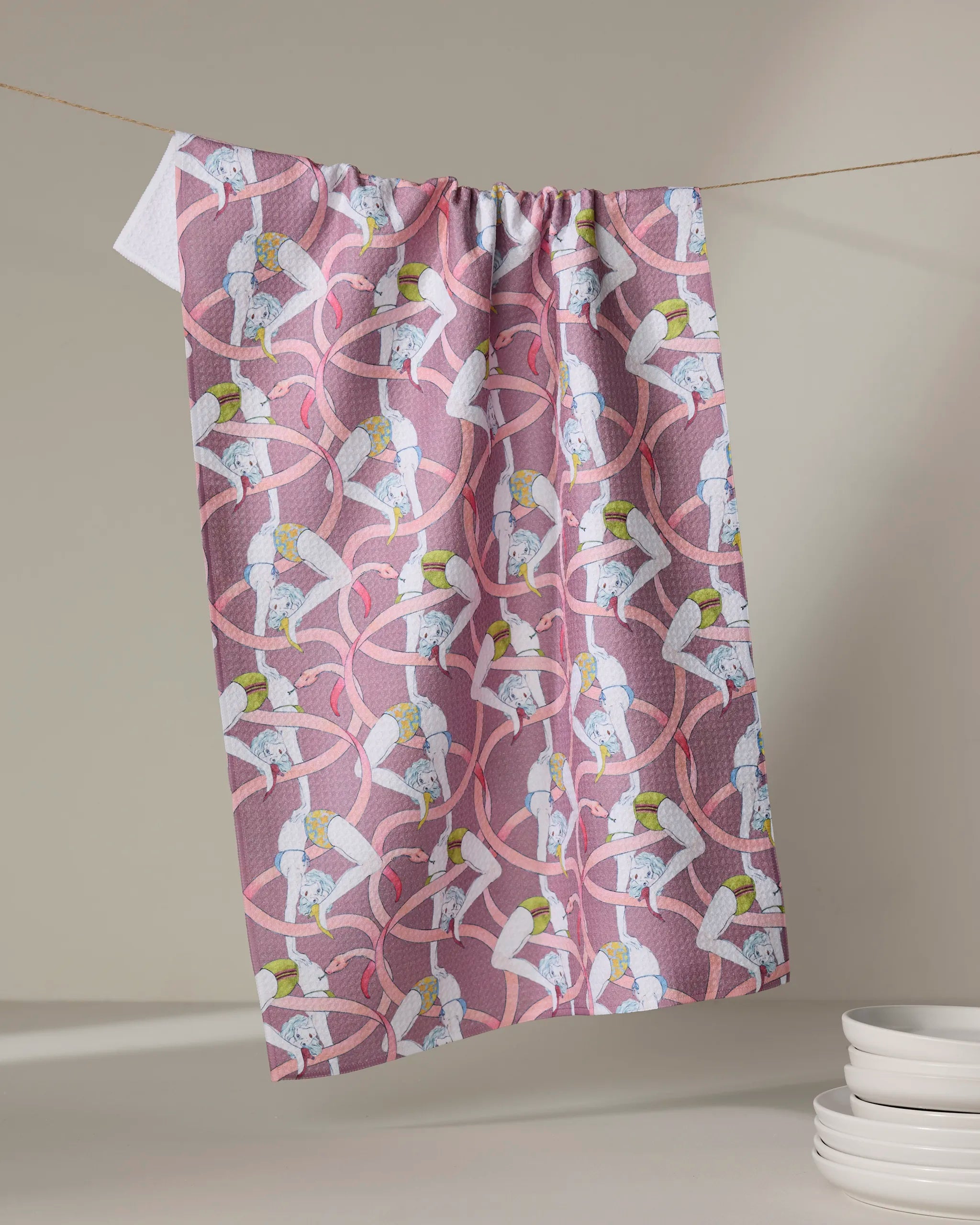 The Circus kitchen towel by Maayan Alper-Swan, featuring eco-friendly fabric and a playful print of women in white leotards and yellow hats with pink ribbons, hangs on a line near stacked white bowls.