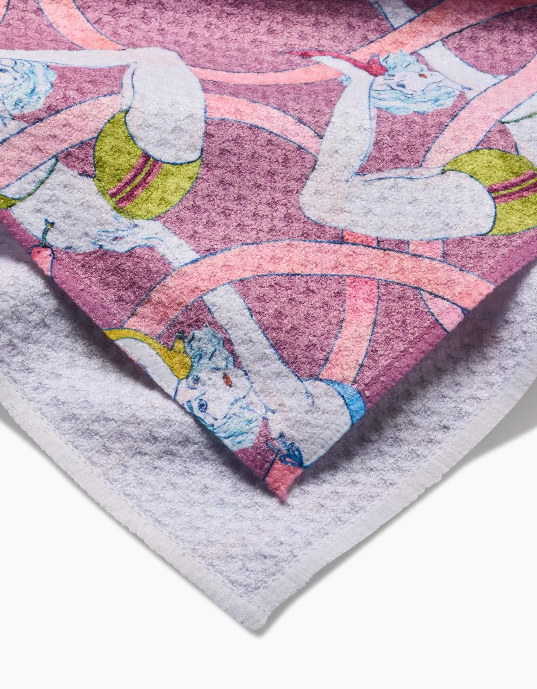 A close-up of the Circus Geoweave™ tea towel by Maayan Alper-Swan, showing illustrated figures in green swimsuits and pink swim caps with pink ribbons on a purple background. The white textured underside is visible. Made from recycled materials.