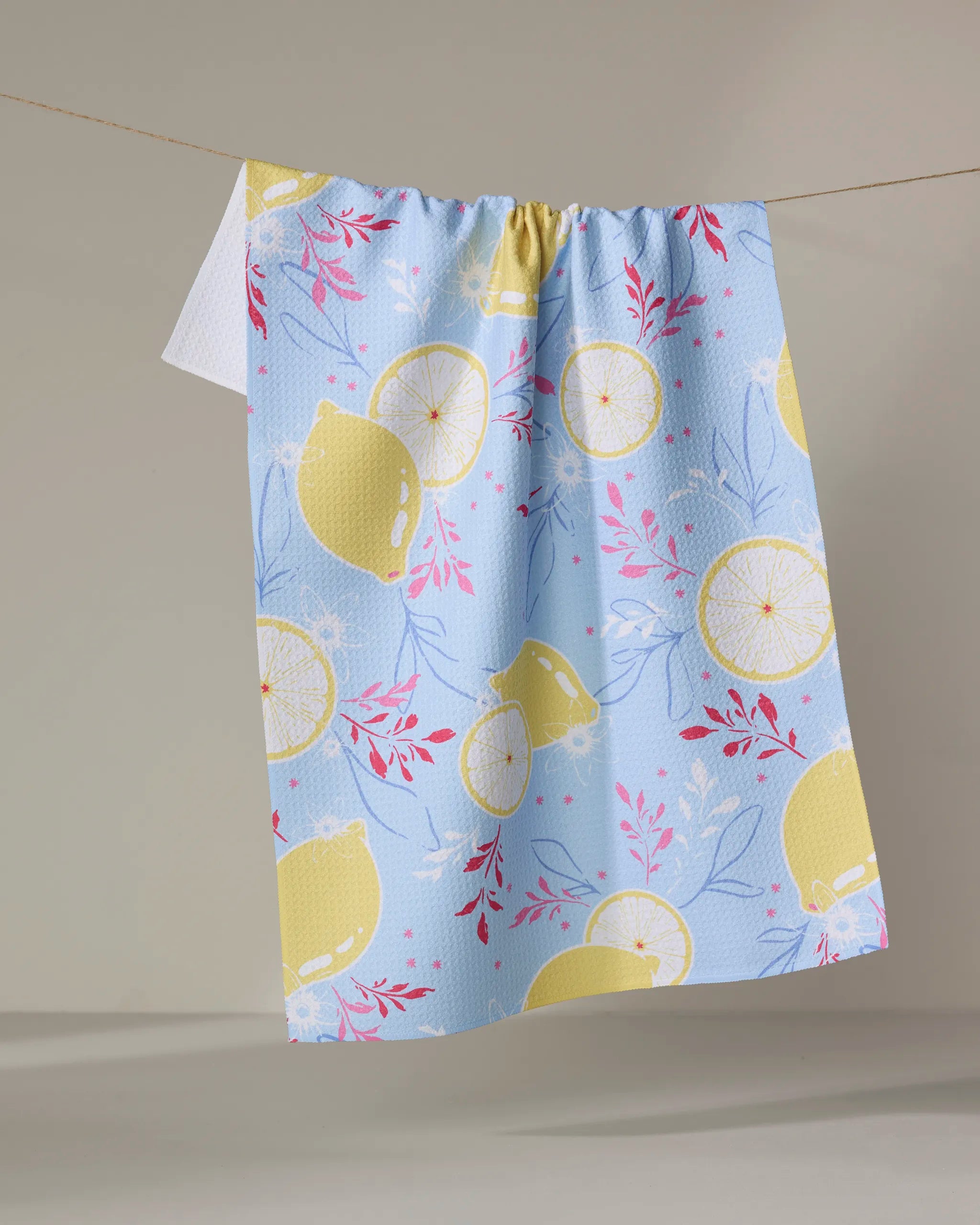 Citrus Bloom by Ashley Celena—a blue recycled kitchen towel with yellow lemons, lemon slices, and pink foliage—hangs on a clothesline against a neutral background.