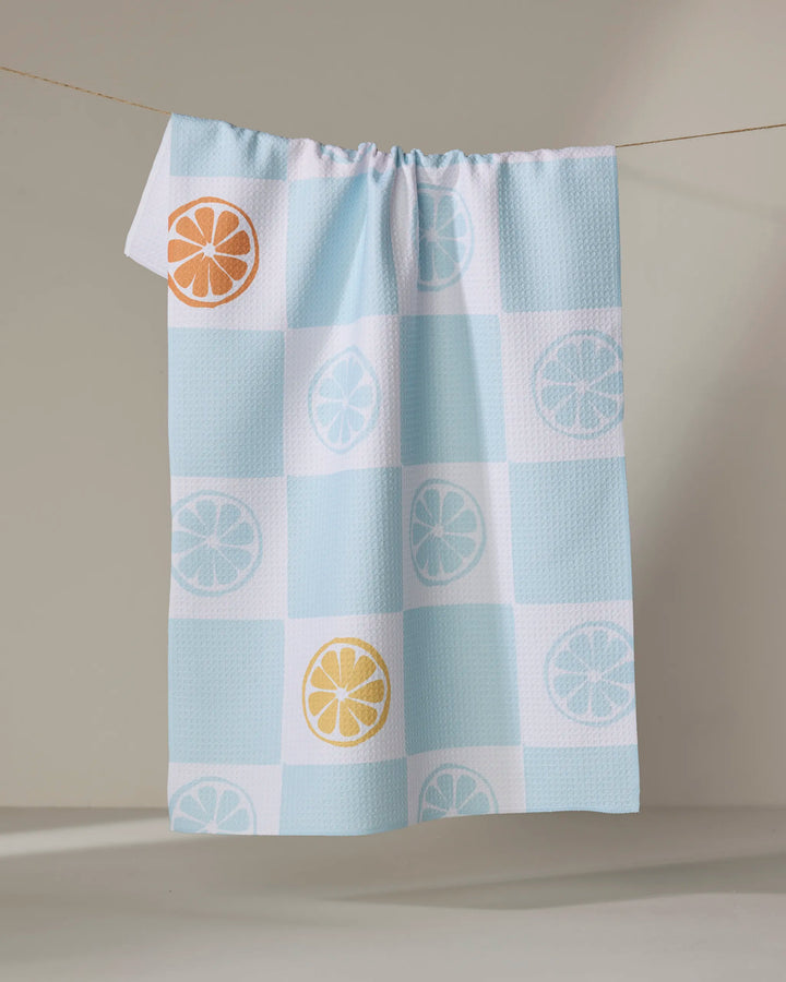 The Citrus Blue tea towel by Janna Sue Design hangs on a clothesline, showcasing blue and white checks with orange, yellow, and light blue citrus slice accents on some squares.