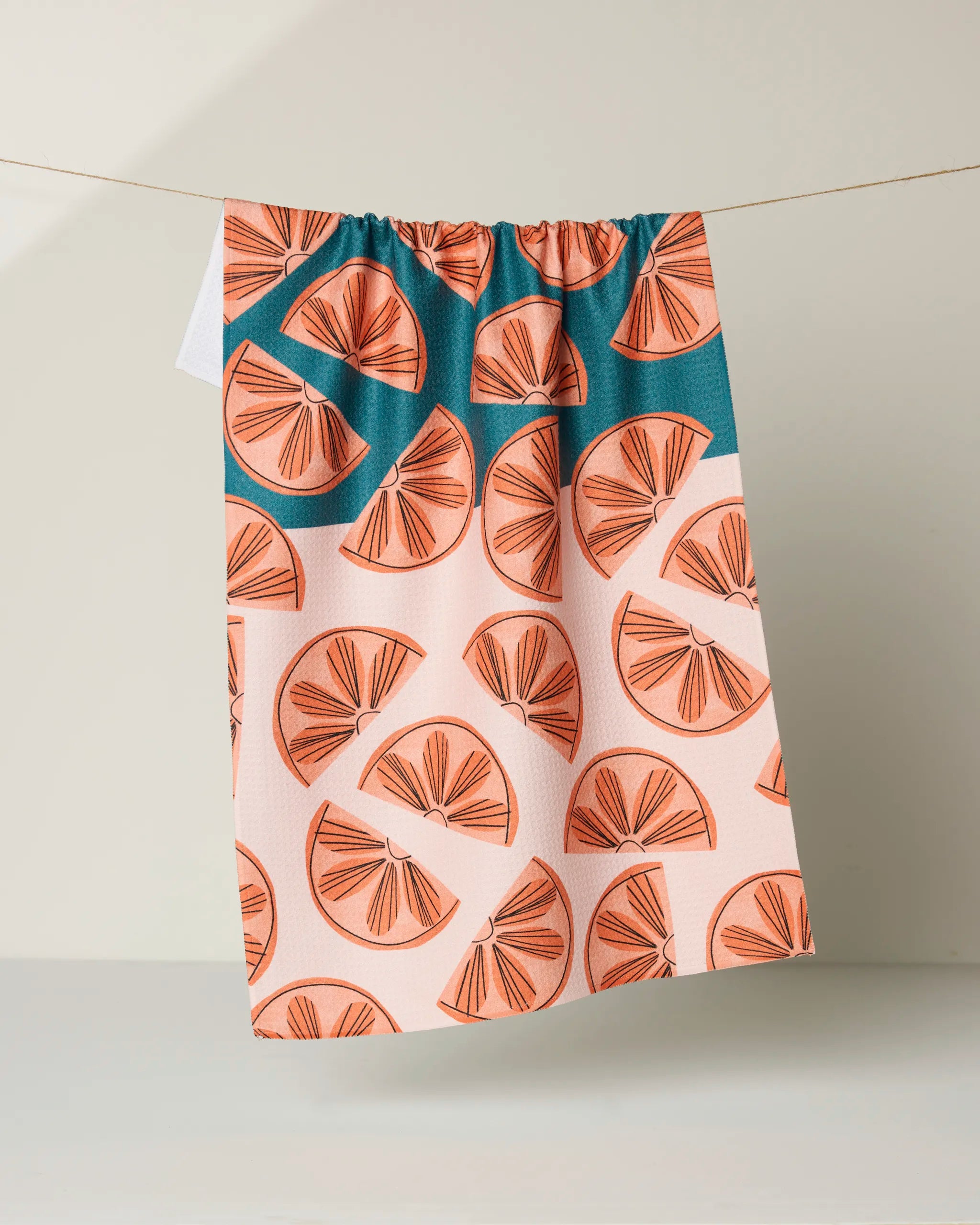 The Louise Pretzel "Citrus Slices" ultra-absorbent kitchen tea towel features an orange slice pattern in peach, coral, and teal, shown hanging on a clothesline against a simple light background.