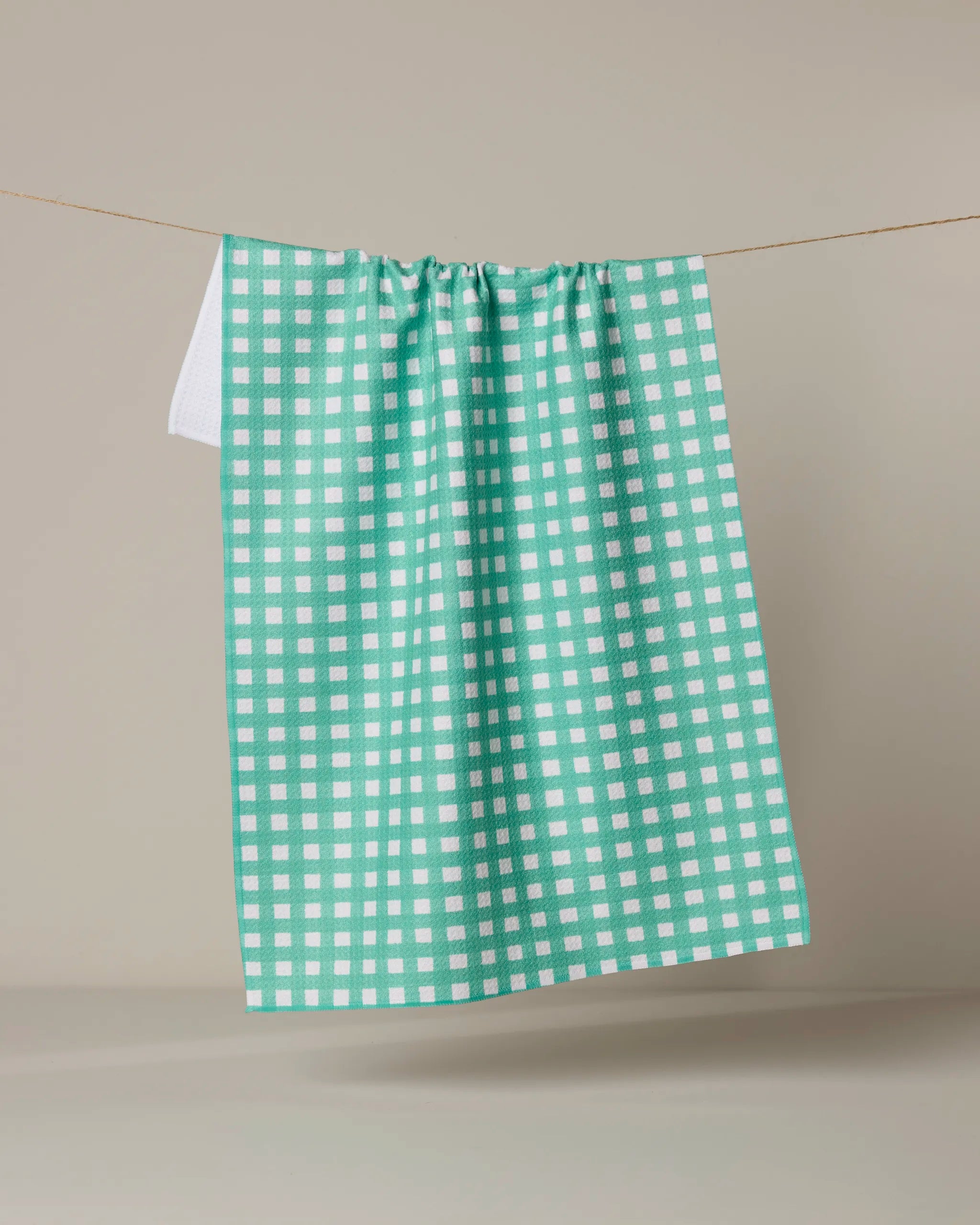 The Claire by Geometry, an ultra-absorbent green and white checkered kitchen tea towel, hangs on a clothesline against a plain beige background.