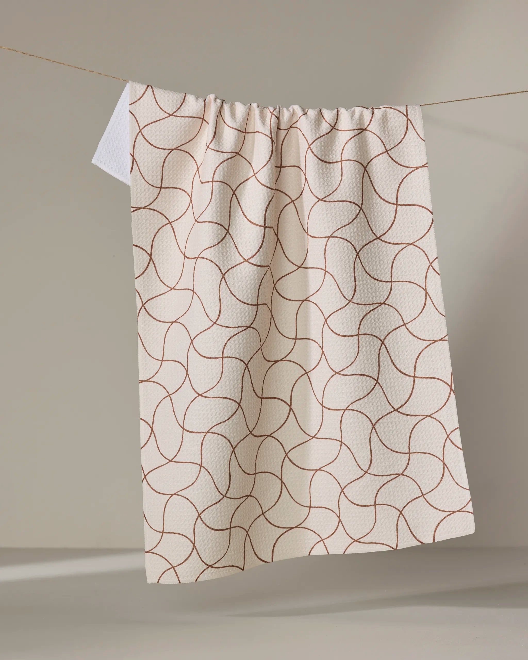 The Clarity tea towel by Gerdadzy, featuring an ultra-absorbent beige fabric with abstract curved brown lines, hangs on a clothesline against a plain, neutral background.