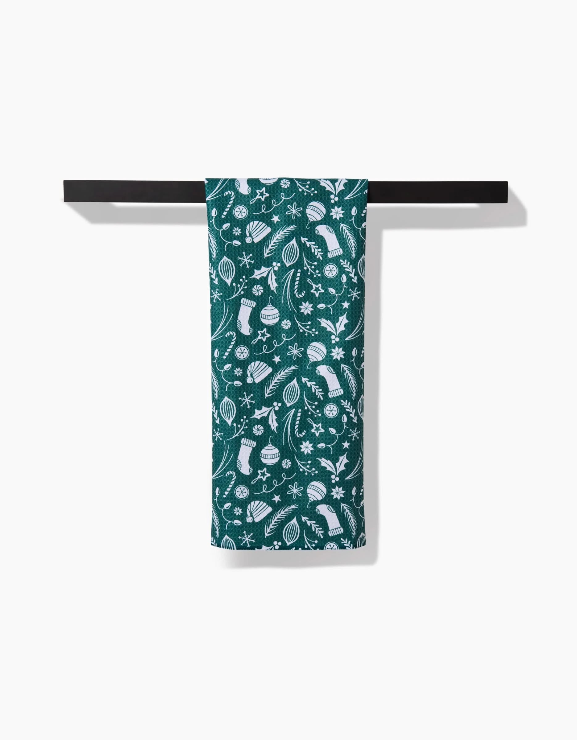 The Sarah Daugherty Classic Christmas Icons Green dish towel, featuring white stockings, ornaments, gifts, and pine branches, hangs over a black towel bar against a white background.