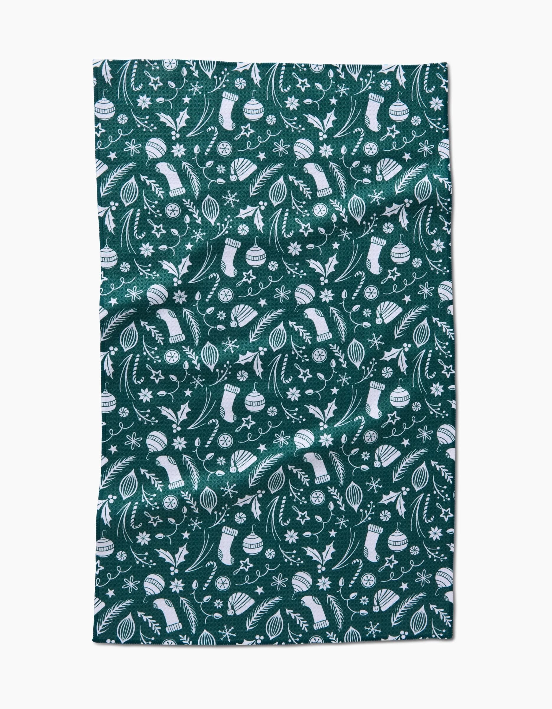 The Classic Christmas Icons Green tea towel by Sarah Daugherty features a white festive pattern with ornaments, stockings, candy canes, holly, snowflakes, stars, and flowers on green. The eco-friendly towel is laid flat with slight wrinkles visible.