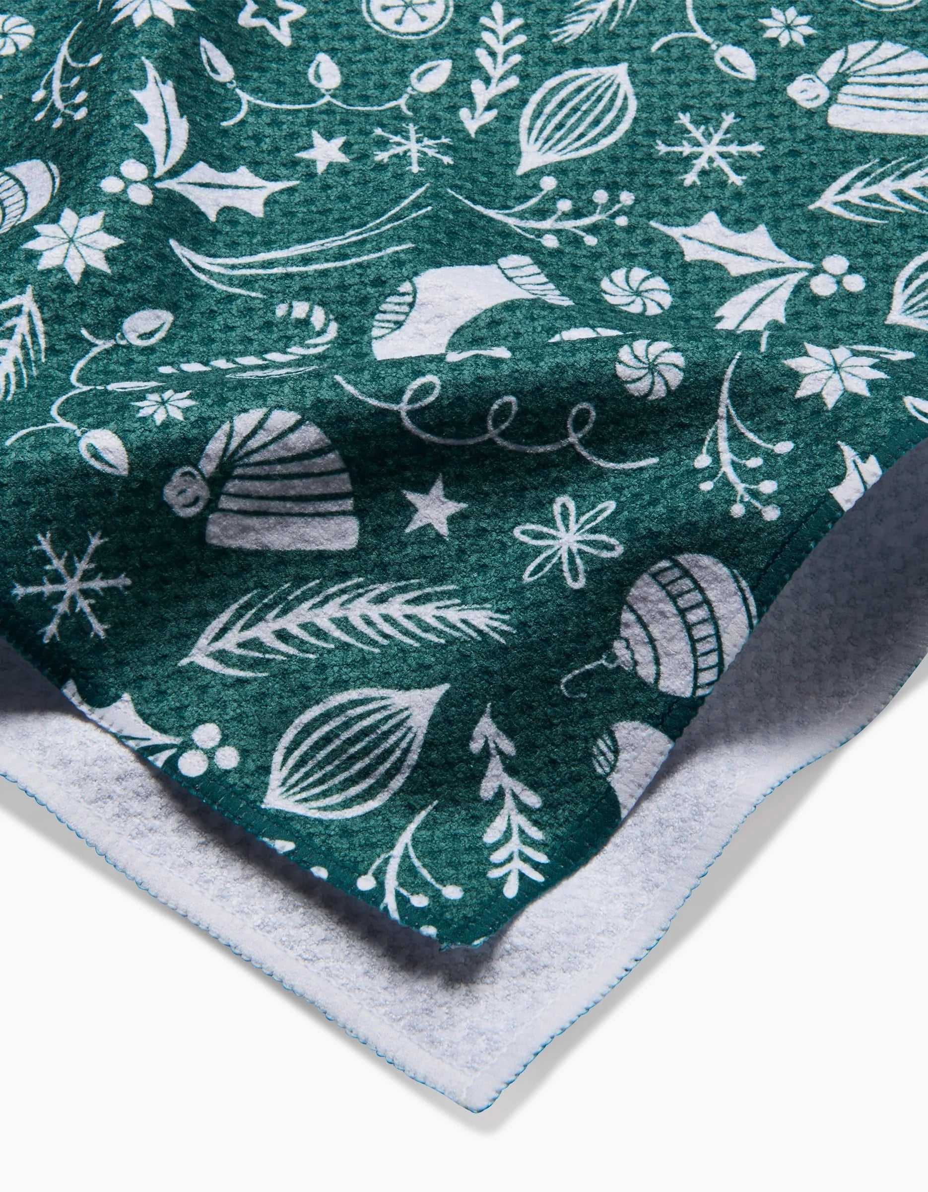 A close-up of the Sarah Daugherty Classic Christmas Icons Green fleece blanket, featuring white ornaments, snowflakes, stars, and candy canes on green. The eco-friendly throw has a soft, textured underside visible at the corner.