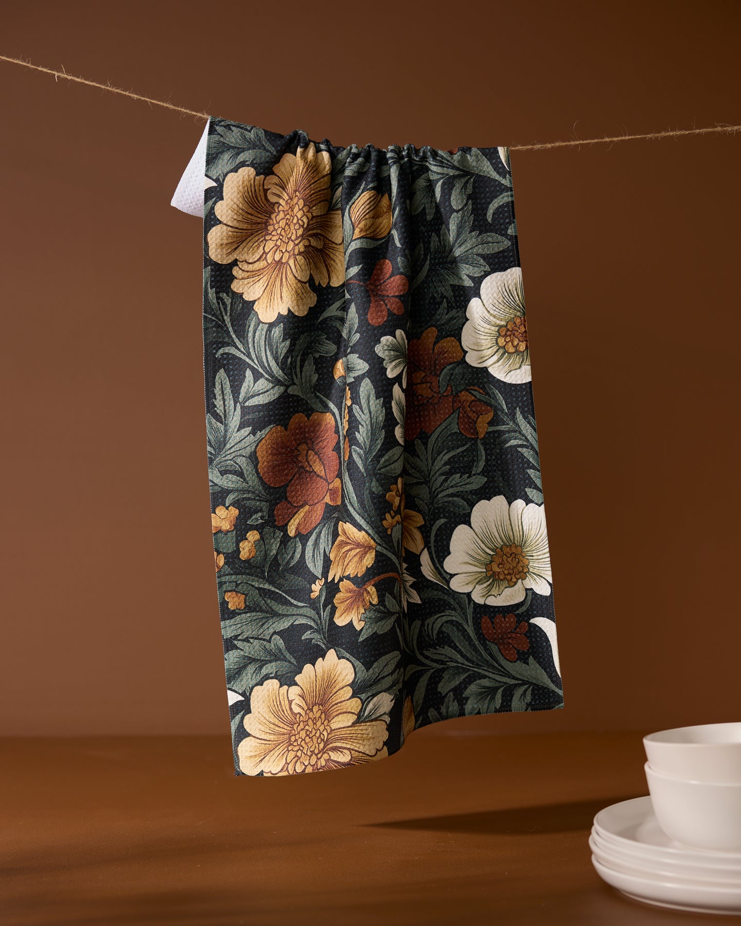 The Geometry Classic Floral eco-friendly kitchen towel, featuring yellow, white, and red flowers, hangs on a string against a brown backdrop with stacked white bowls and plates on the right.