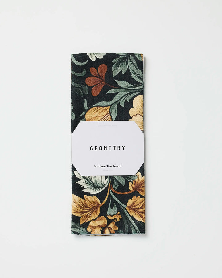 A folded eco-friendly Geometry Kitchen Tea Towel in the Classic Floral dark print with yellow, green, and rust accents, wrapped in a white label displaying the brand name. Set against a white background.