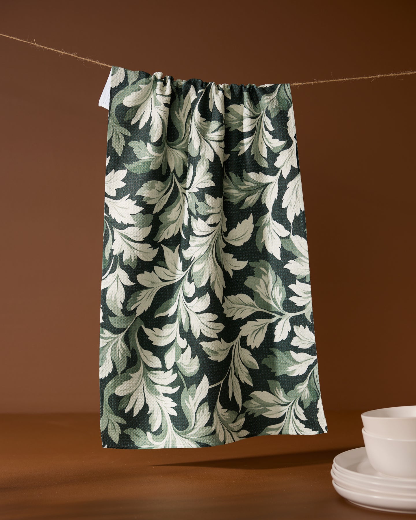 The Geometry Classic Foliage kitchen towel, featuring a white leafy design on dark green and made from recycled materials, hangs from a clothesline against a brown background. Below, white dishes are neatly stacked.