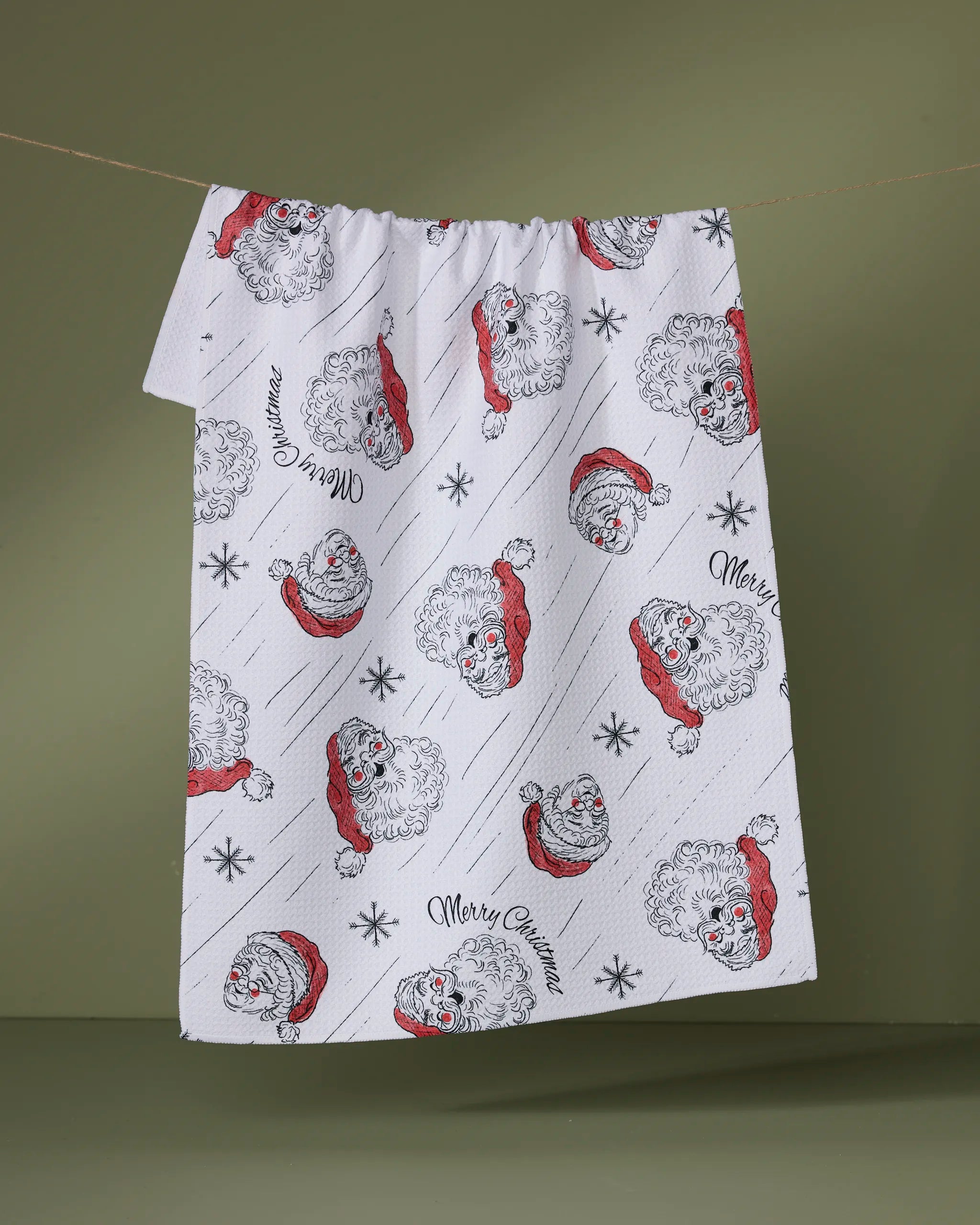 The Claus Family Christmas tea towel by Geometry features Santa faces, snowflakes, and "Merry Christmas" in black script, displayed on an olive-green background.