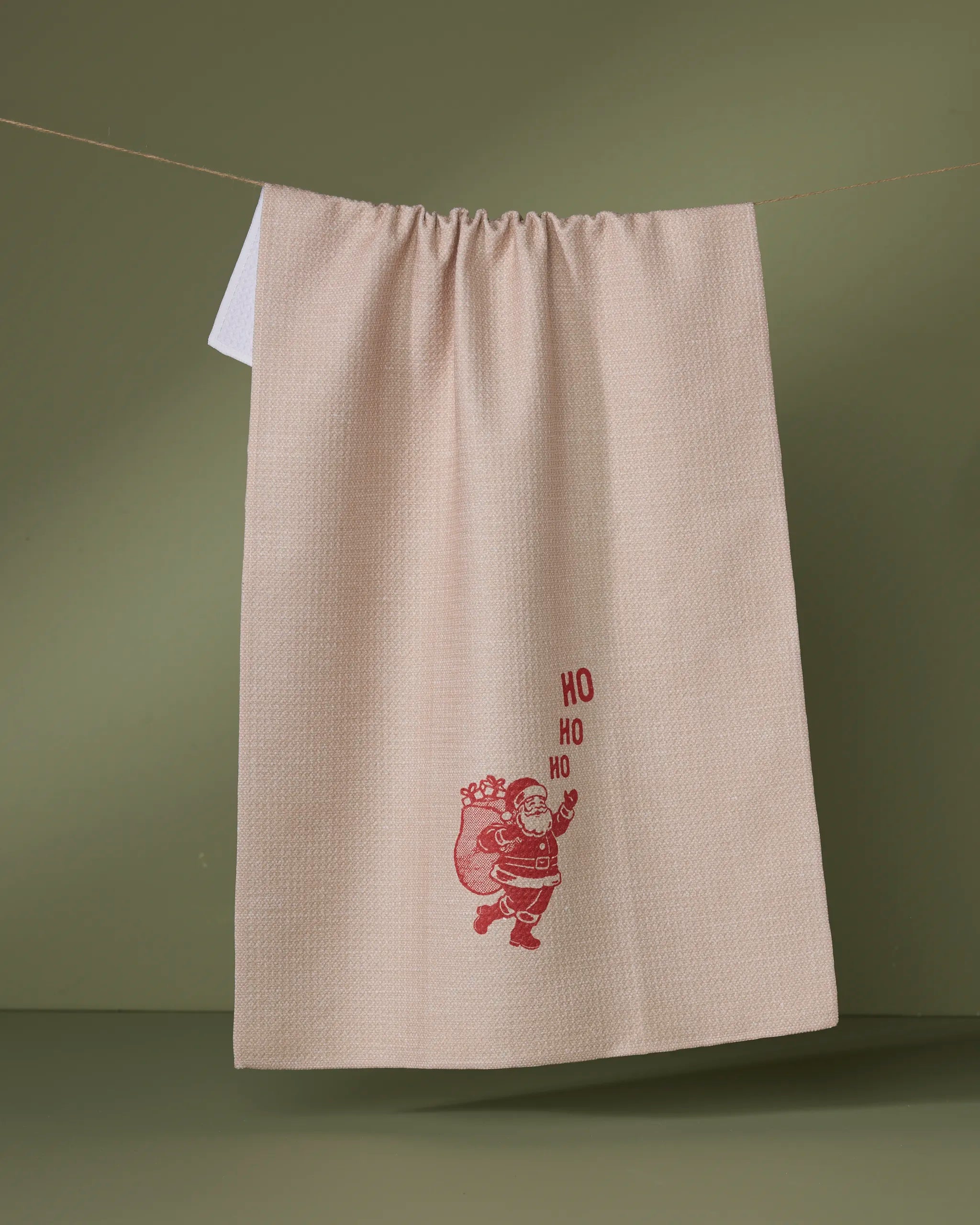 The Geometry Clausco recycled kitchen towel in beige hangs on a line against a green background, showcasing a red Santa Claus with a sack and the phrase "HO HO HO.