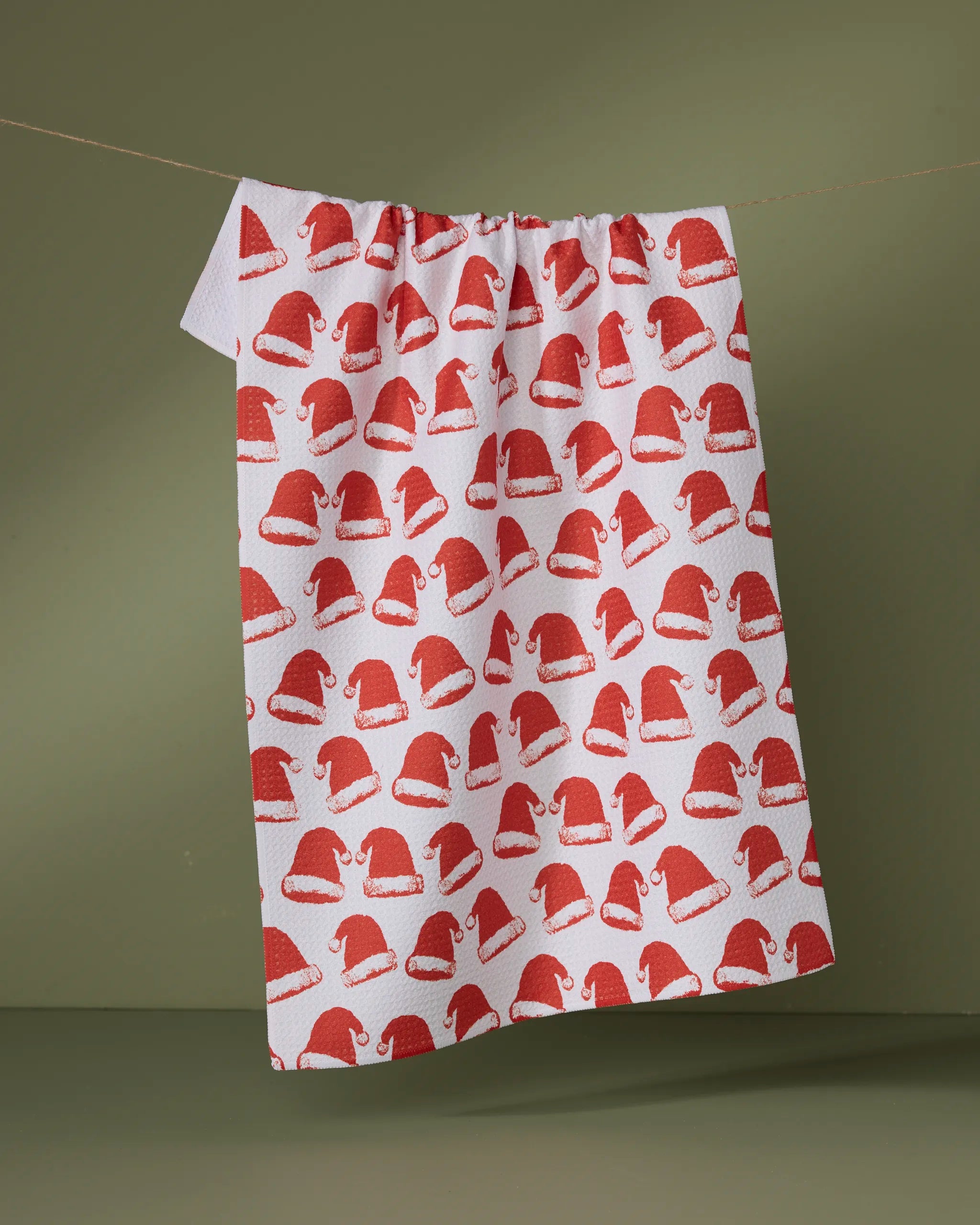 The Clausen tea towel by Geometry hangs on a line against an olive-green background. Made from ultra-absorbent recycled materials, it features a festive red Santa hat pattern and is both eco-friendly and stylish.