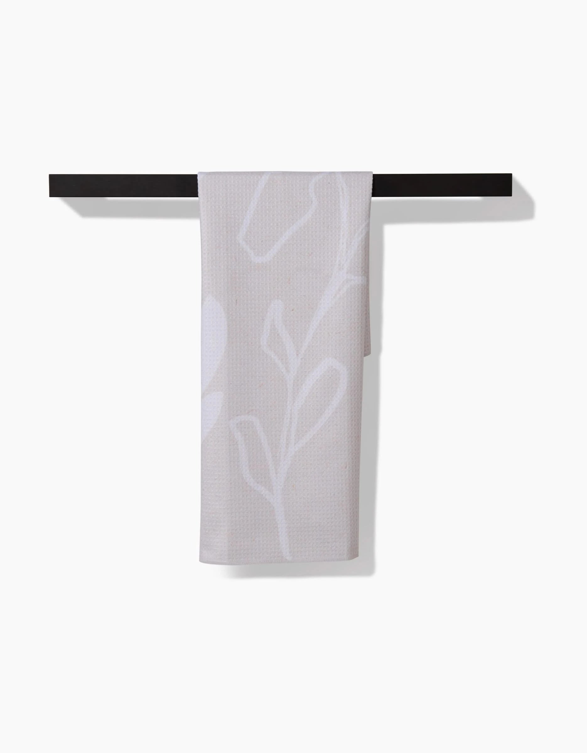 The GEOMETRY Clean Tea Towel, made from recycled materials and featuring a subtle white leaf pattern in beige, is draped over a black bar against a white background.