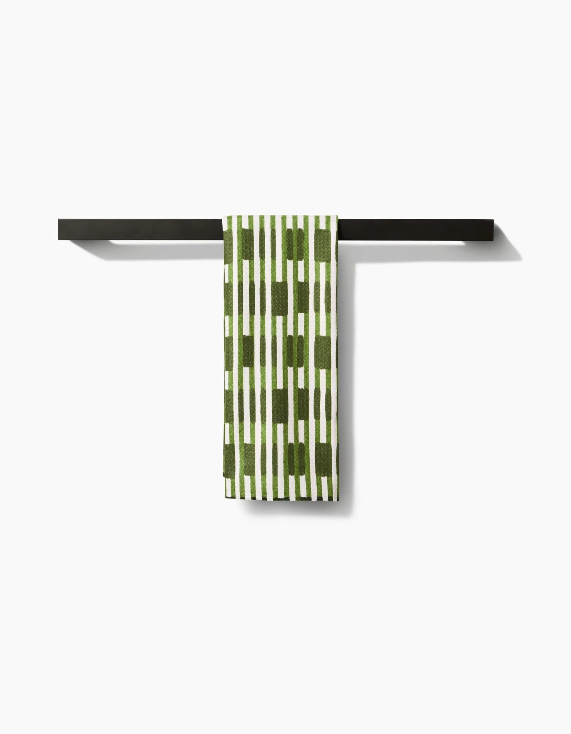 The GEOMETRY Clover Stripe recycled kitchen towel, featuring a green and white pattern, hangs neatly over a sleek black horizontal towel bar against a plain white background.