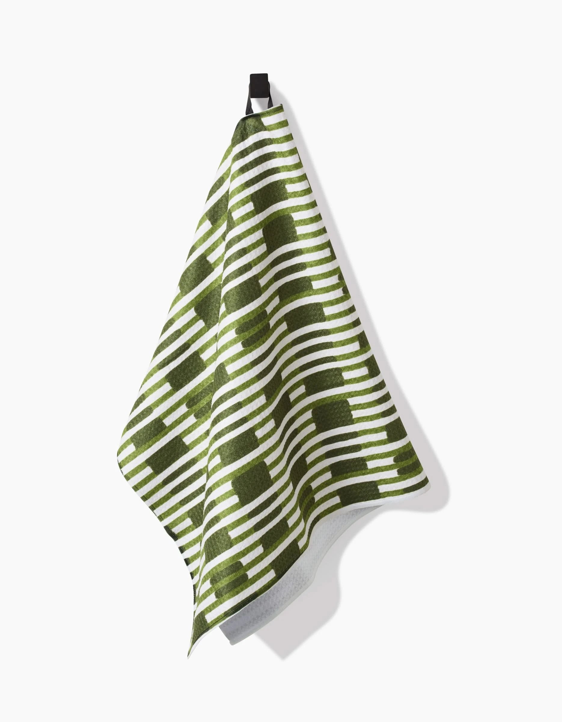 The Clover Stripe kitchen towel by GEOMETRY, featuring a green and white geometric pattern, hangs from a black hook against a white background.