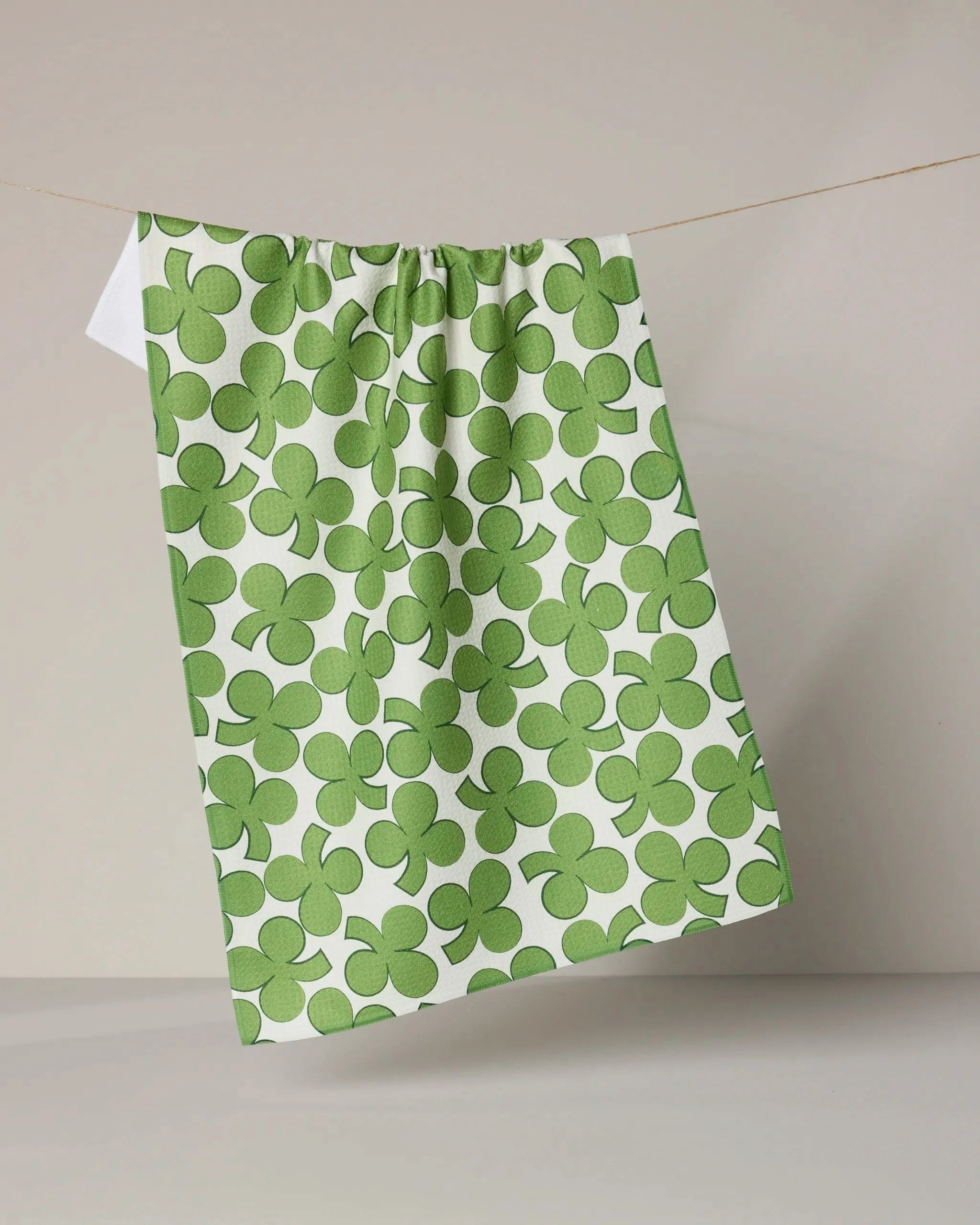 The Geometry Clover Toss dish towel, featuring a green clover pattern, hangs on a clothesline against a plain light gray background.