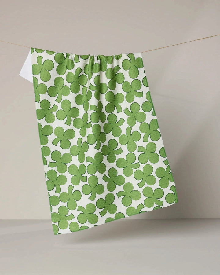 The Geometry Clover Toss dish towel, featuring a green clover pattern, hangs on a clothesline against a plain light gray background.