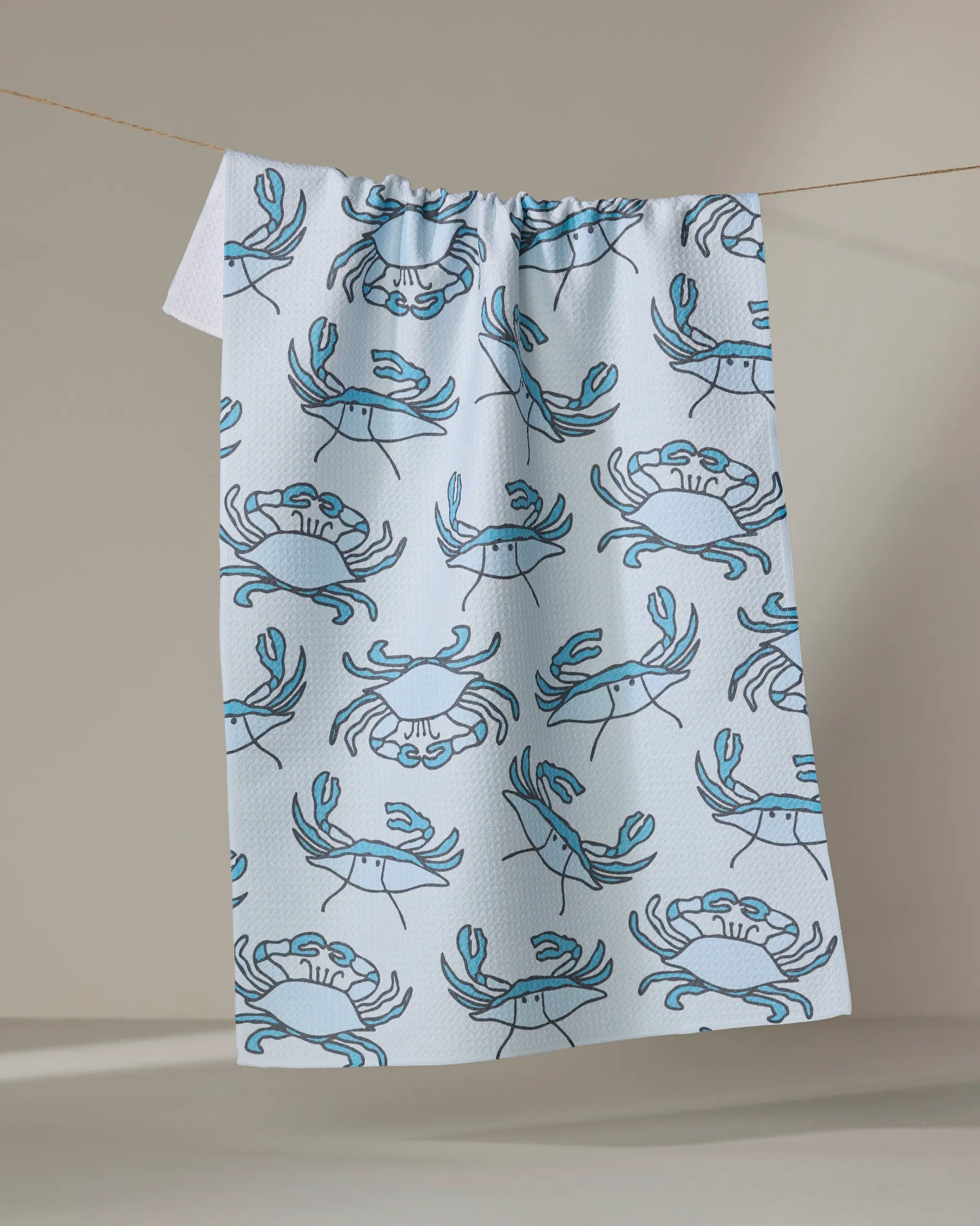The Mindfulnice Coastal Crew tea towel, featuring a quick-drying white fabric with a repeating blue crab pattern, hangs on a line against a neutral background.