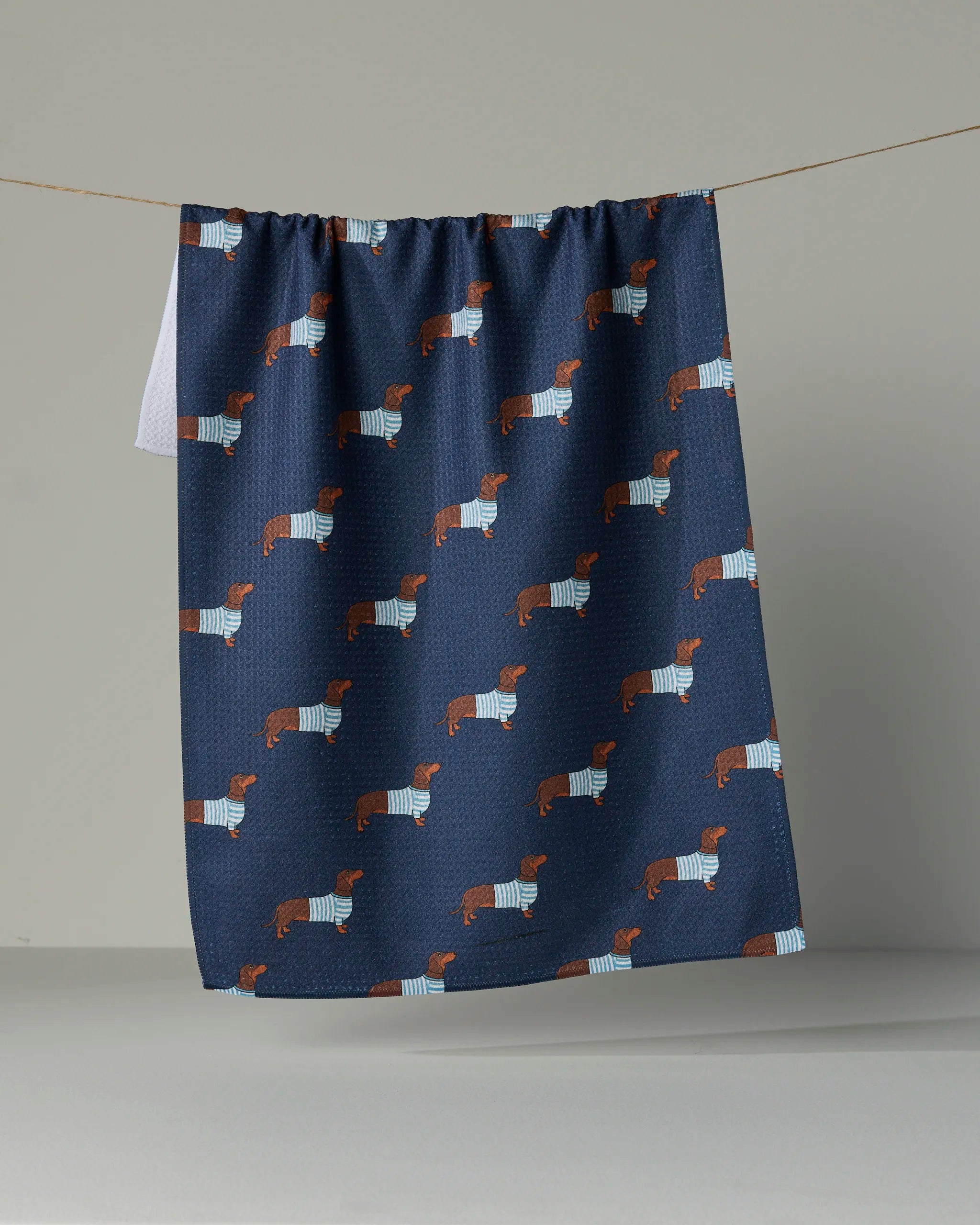 The Geometry Coastal Dachshund kitchen towel, crafted from recycled materials, displays a navy blue design with a repeating pattern of dachshund dogs in blue and white striped sweaters, shown hanging on a line against a plain background.