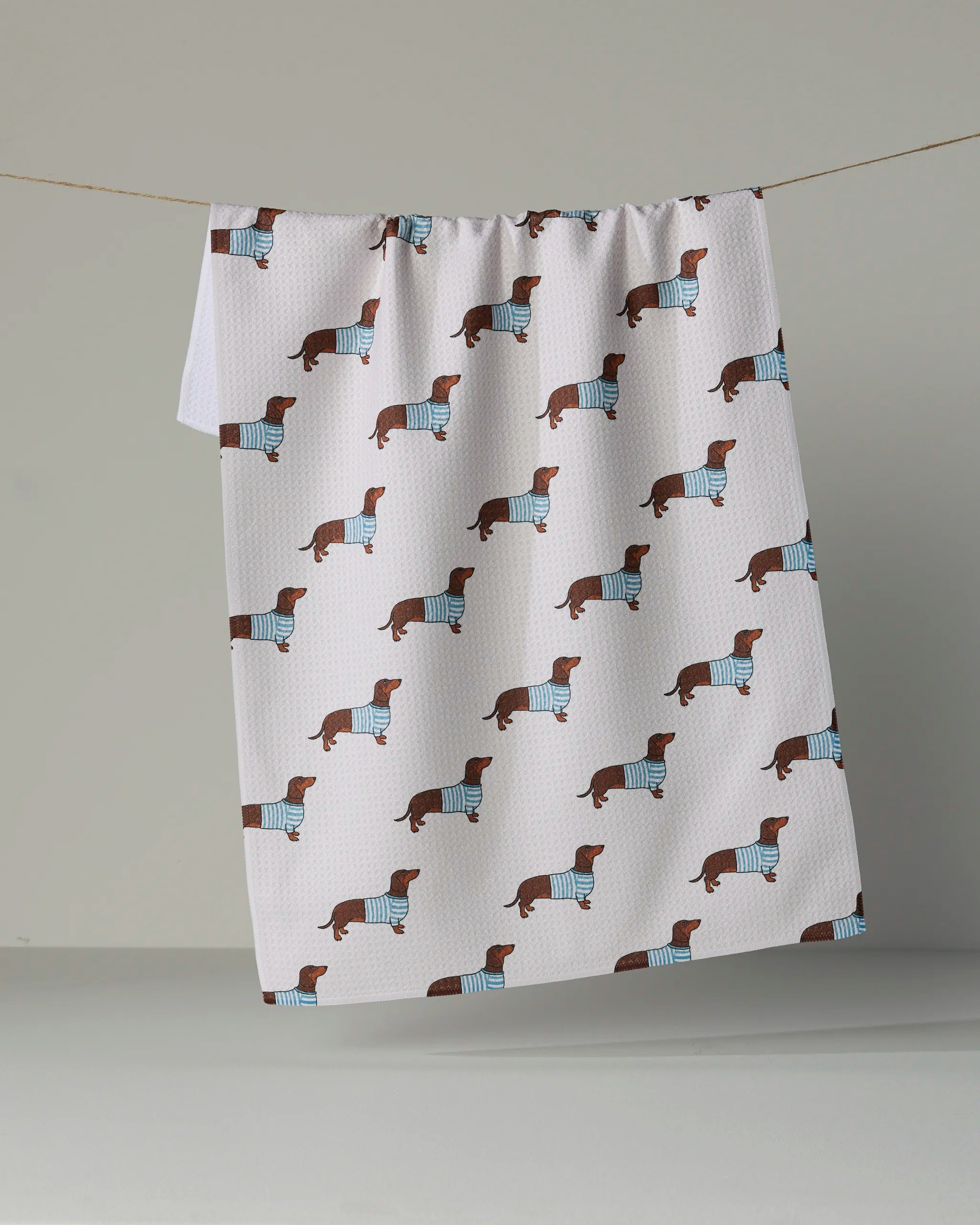 The Geometry “Coastal Dachshund” kitchen towel, crafted from recycled materials and adorned with a repeating pattern of dachshunds in blue and white striped sweaters, hangs on a clothesline against a neutral backdrop.