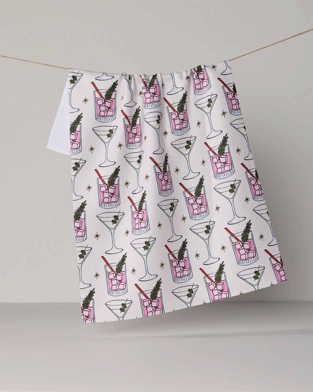 The Geometry “Cocktail Countdown” tea towel features a stylish pattern of martini glasses and cocktail tumblers accented with olives and herbs, hanging on a line against a plain backdrop. Made from recycled materials, this ultra-absorbent tea towel brings both sustainability and flair to your kitchen décor.