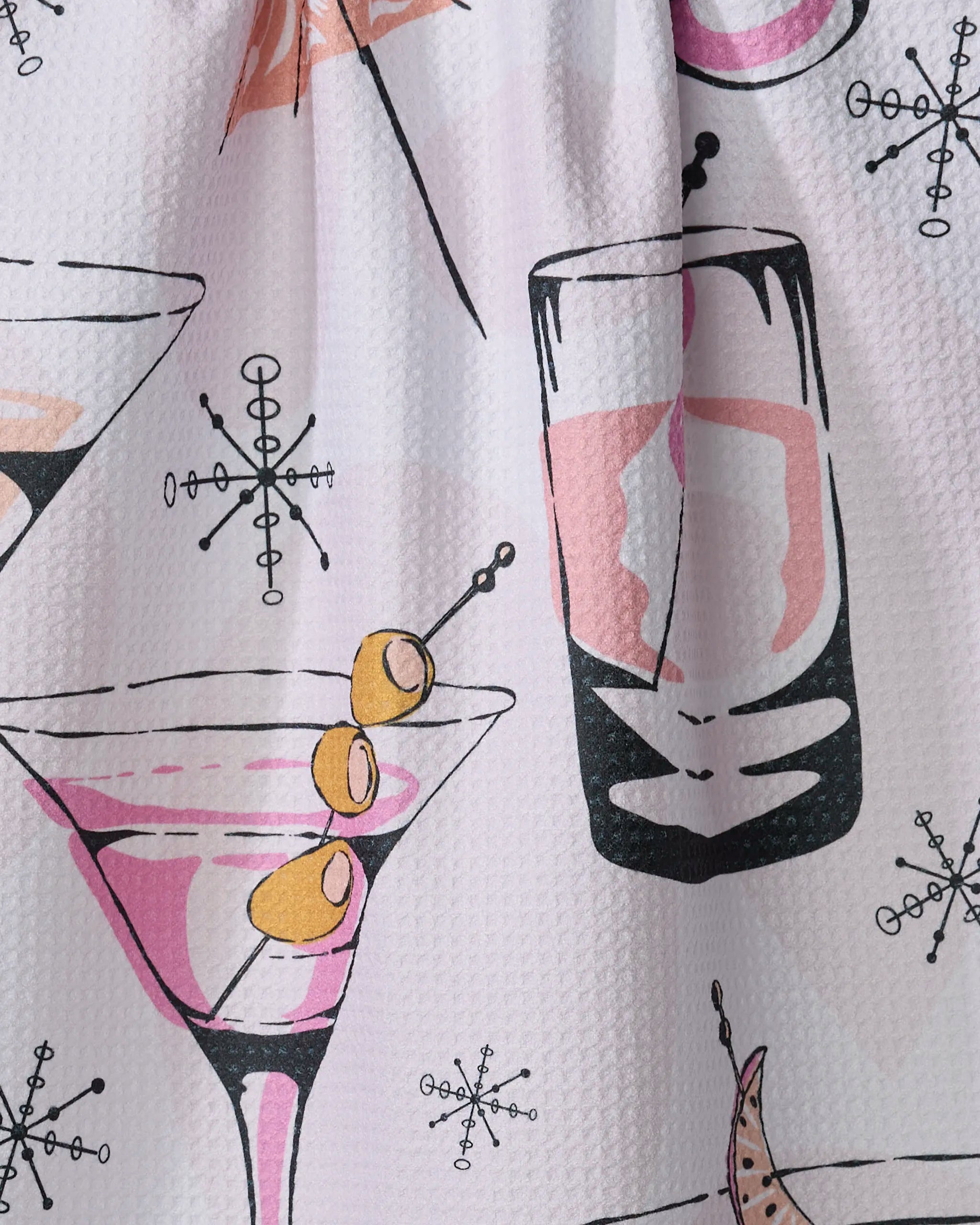 A close-up of Julianne Haness's "Cocktail Hour" tea towel, ultra-absorbent and made from recycled materials, features a retro print with martini glasses, pink drinks, and atomic starbursts on a white textured background.
