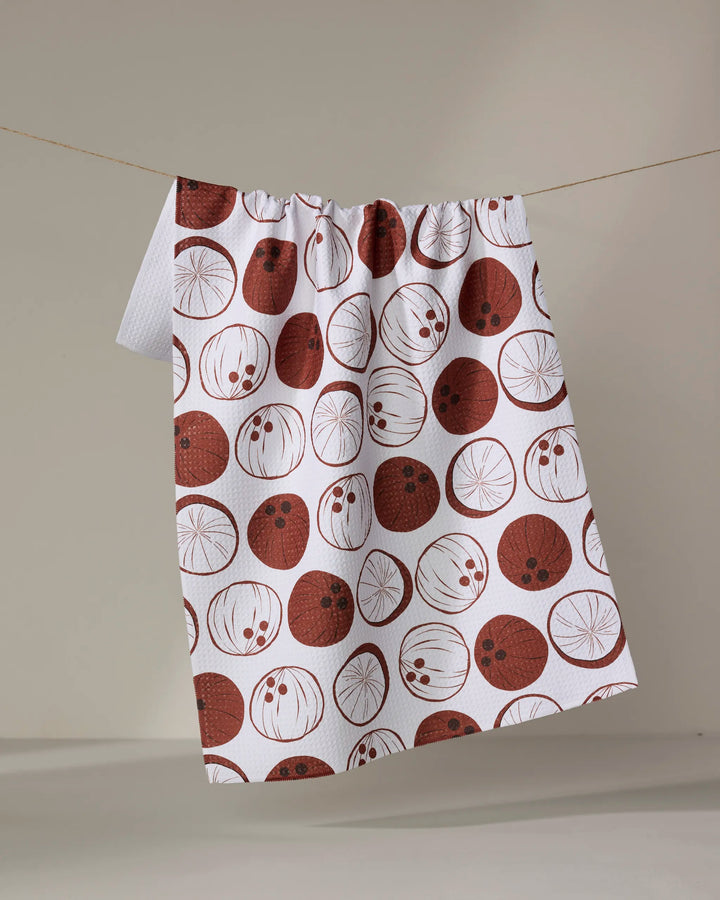 The "Coconutz About You" tea towel by Sara Mari features red and brown circular fruit slice patterns on white, crafted from ultra-absorbent fabric, and hangs on a clothesline against a plain, neutral background.