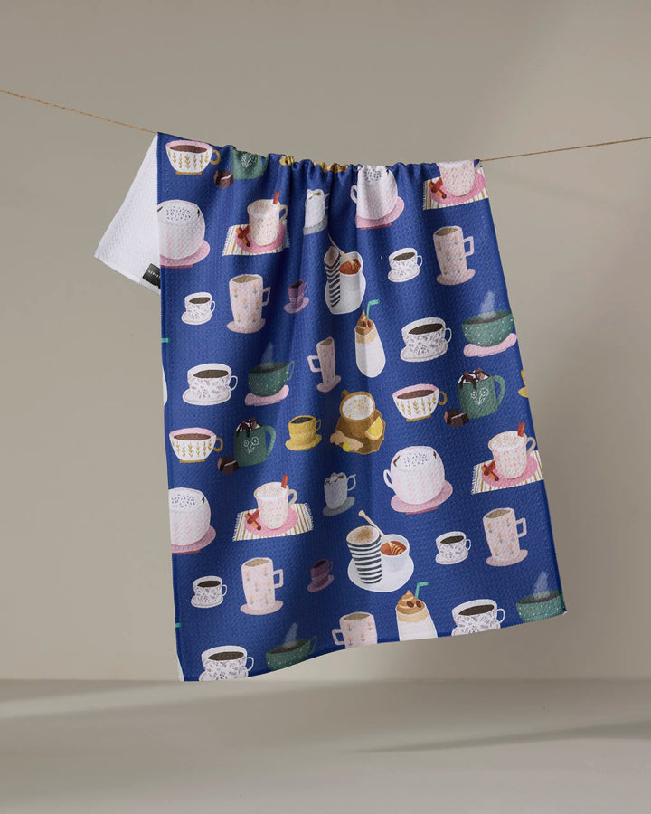 The Coffee Drinks kitchen towel by Cottonflower Studio is blue, made from recycled materials, and features colorful mugs, cups, teapots, and desserts hanging on a clothesline against a neutral background.