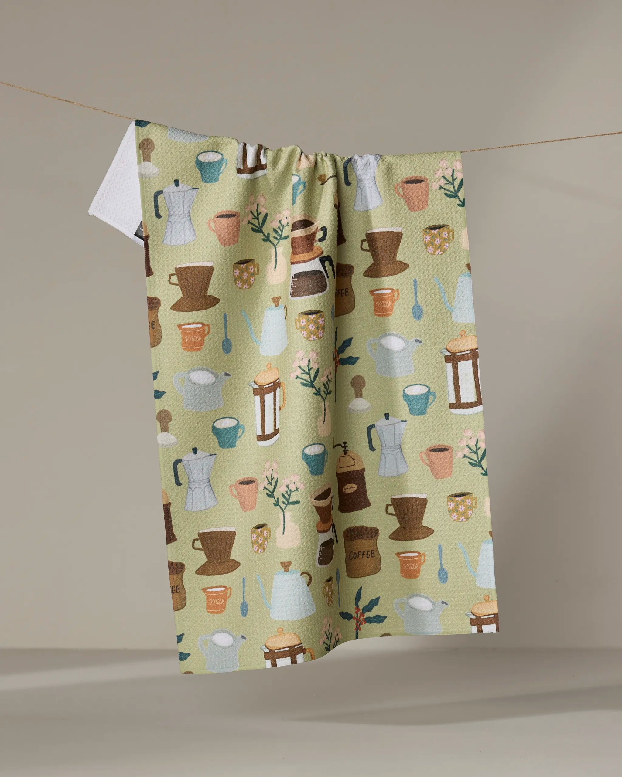 The "Coffee Lover" kitchen towel by Cottonflower Studio features pastel coffee pots, mugs, cups, spoons, jars, and plants—crafted from recycled materials and shown hanging on a clothesline against a neutral backdrop.
