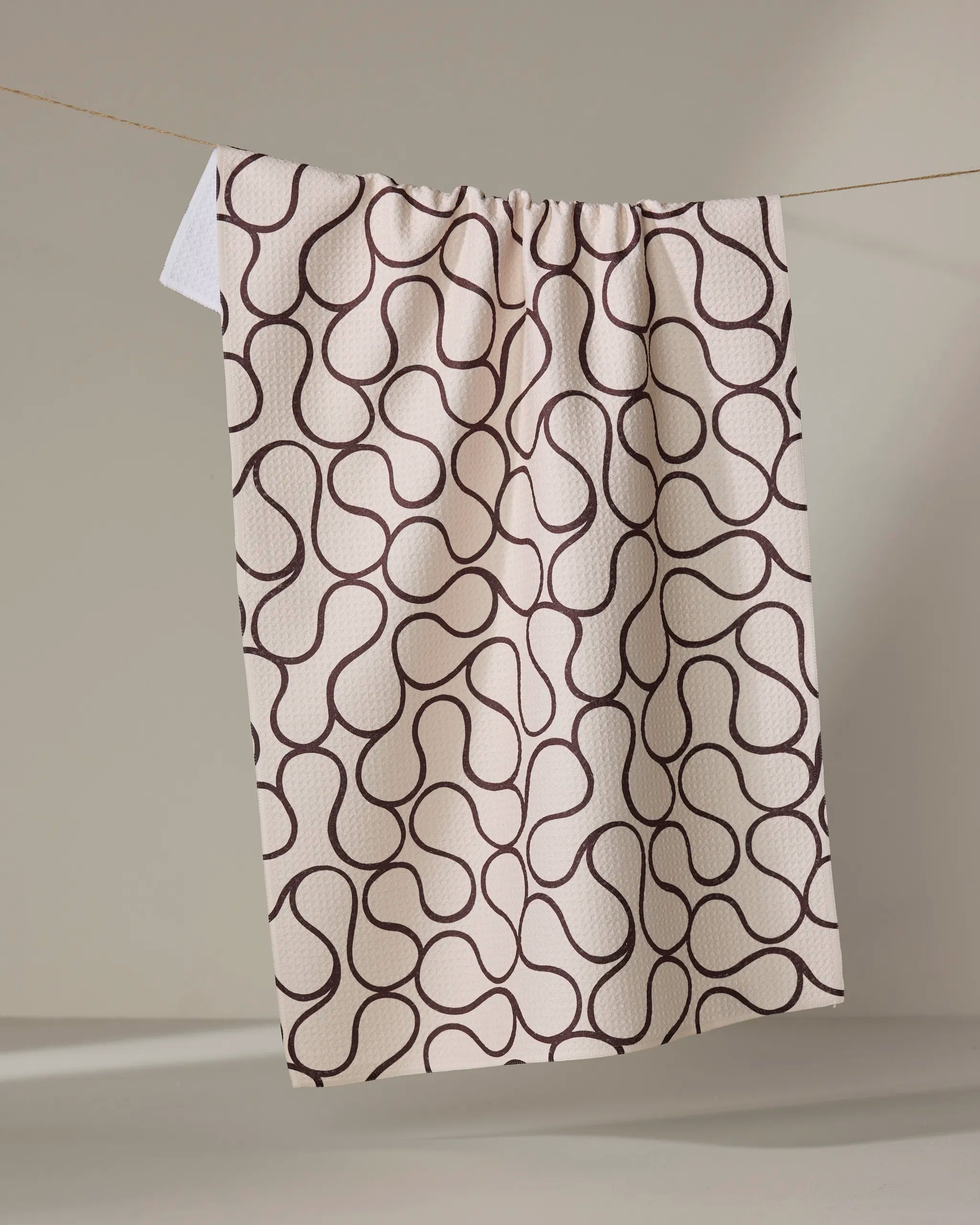 The Gerdadzy Coffee Swirl towel features a cream color with a repeating wavy brown geometric pattern and hangs on a thin line against a neutral background.
