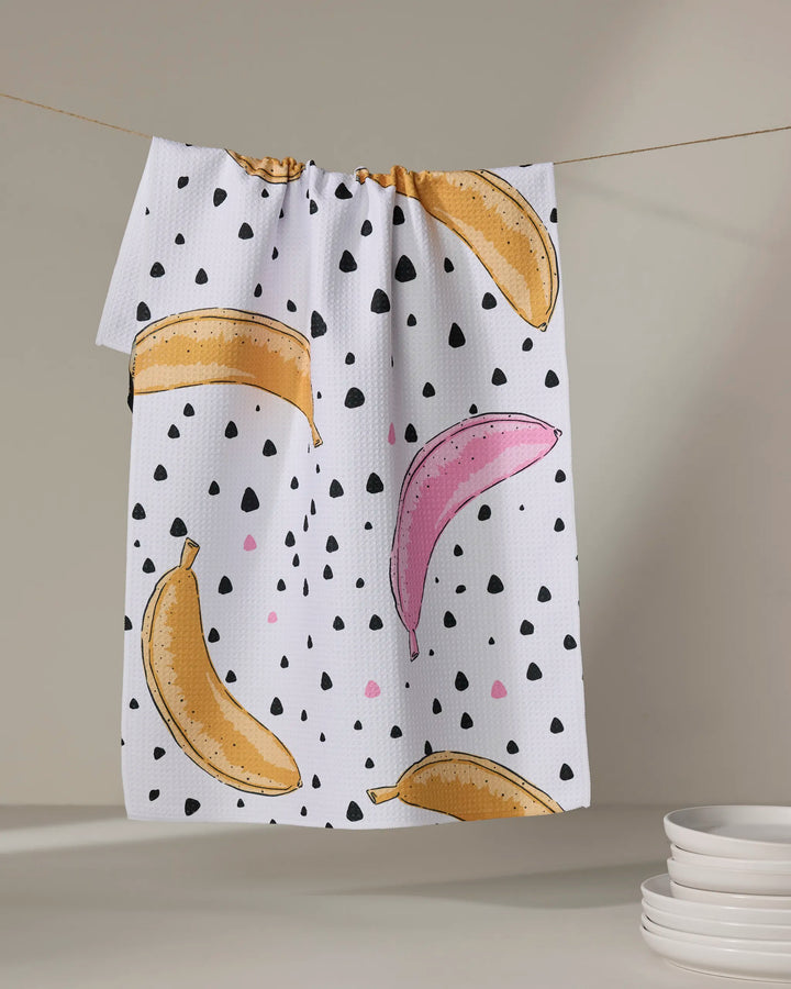 The Julianne Haness Color Pop Bananas tea towel, featuring yellow and pink bananas with black and pink dots, hangs on a clothesline beside a stack of white plates.